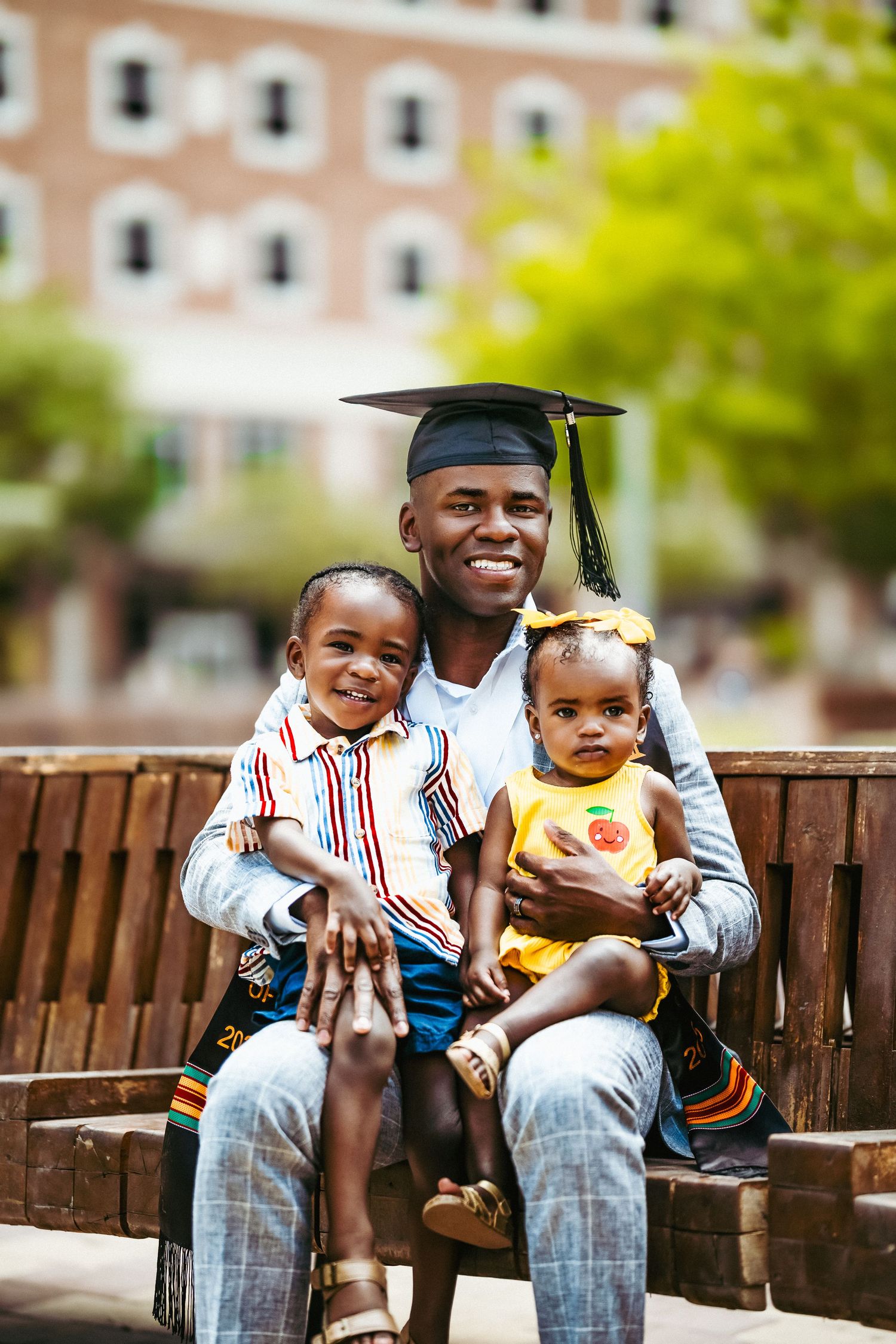 Graduate in cap and gown sits on bench with two young children on a sunny day near brick building.