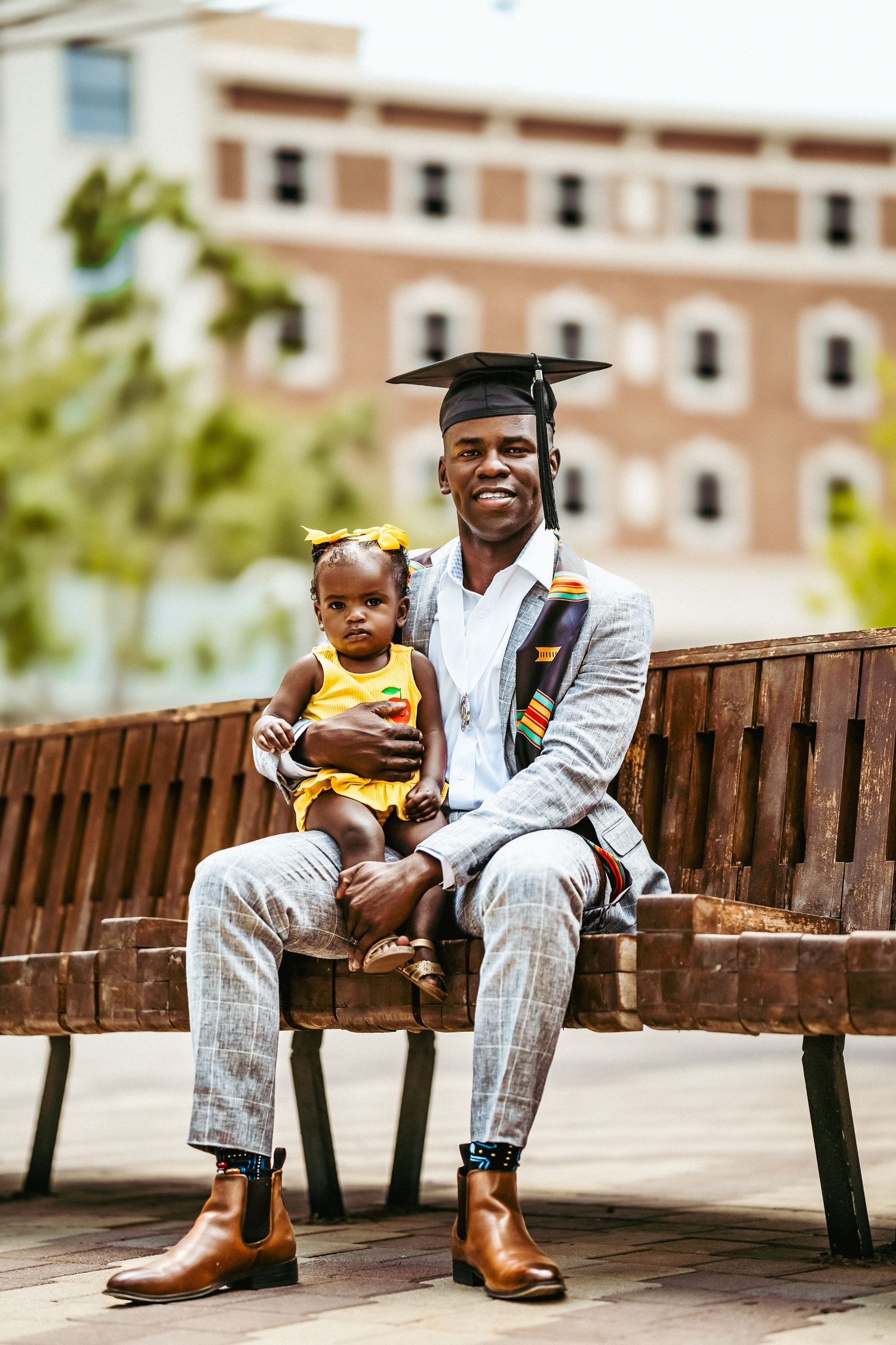 A graduate in a light suit sits with a child in yellow on a wooden bench in an outdoor setting.