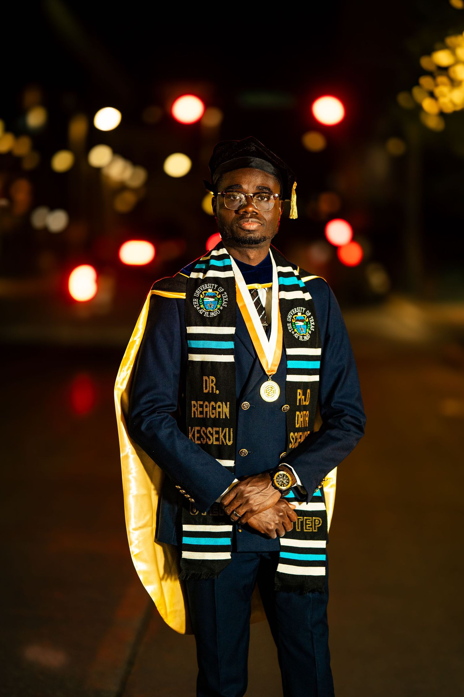 Graduate wearing navy blue academic regalia with teal stole and yellow accents poses at night with bokeh lights in background.