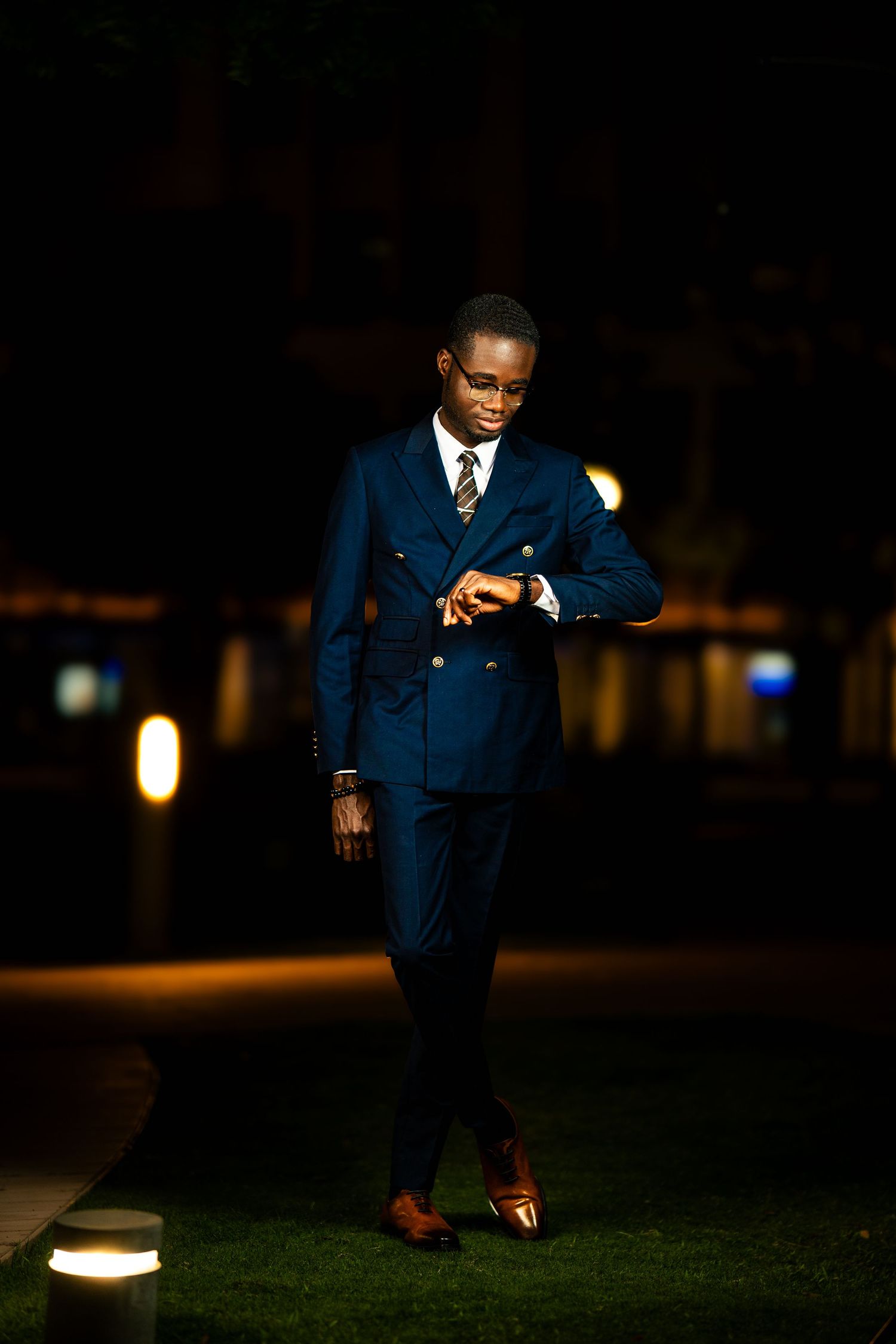 A person in a navy blue suit poses outdoors at night on grass near illuminated lighting fixtures.