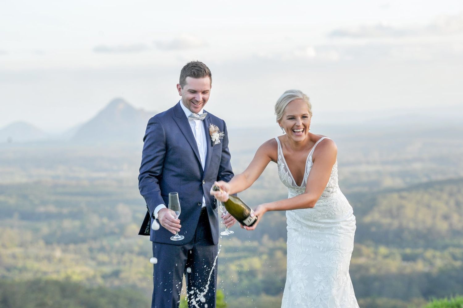 Joyful bride pops champagne bottle with groom against scenic mountain landscape backdrop.
