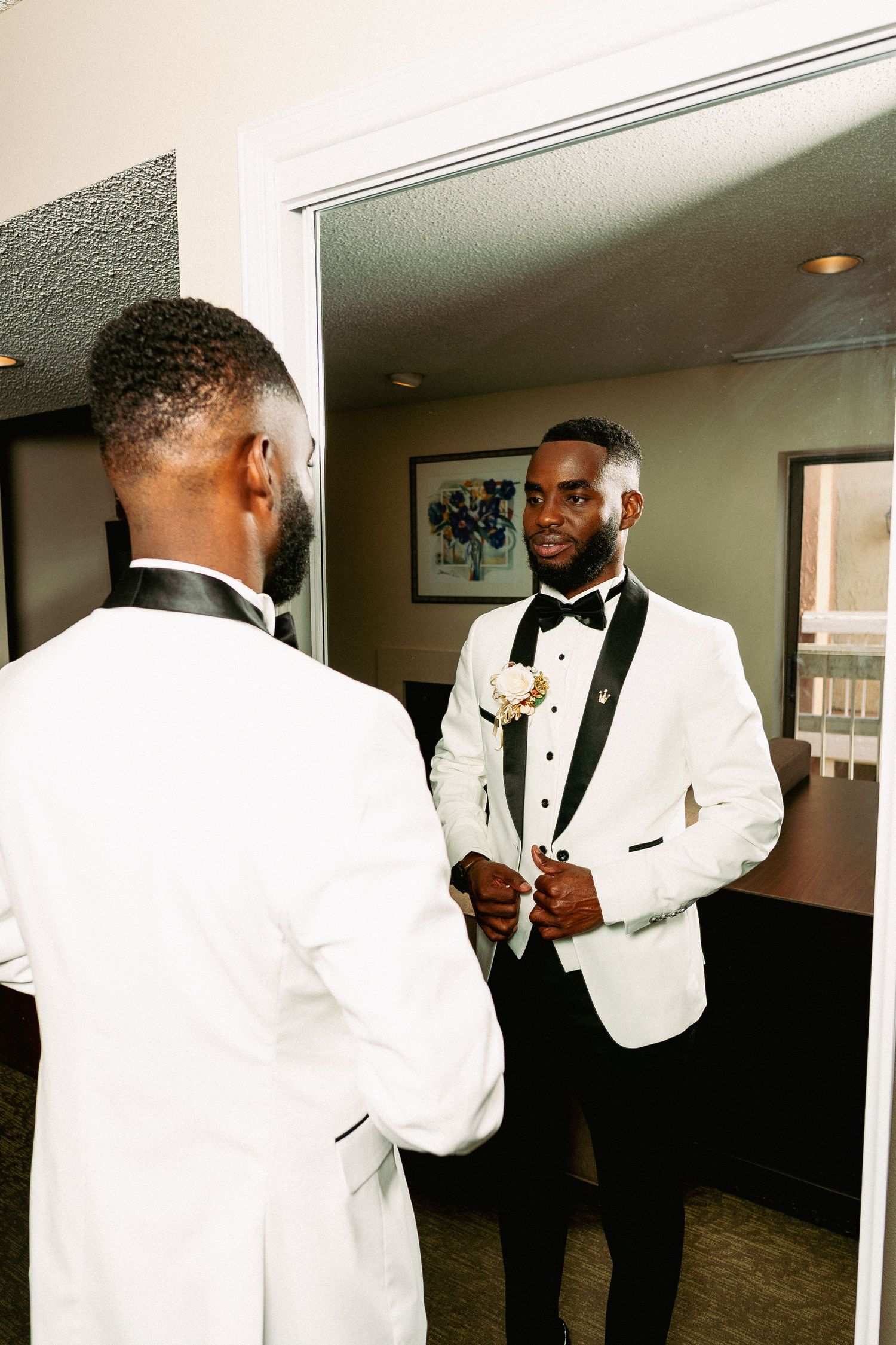 A groom in a white tuxedo jacket and black bow tie adjusts his outfit while looking in a mirror.