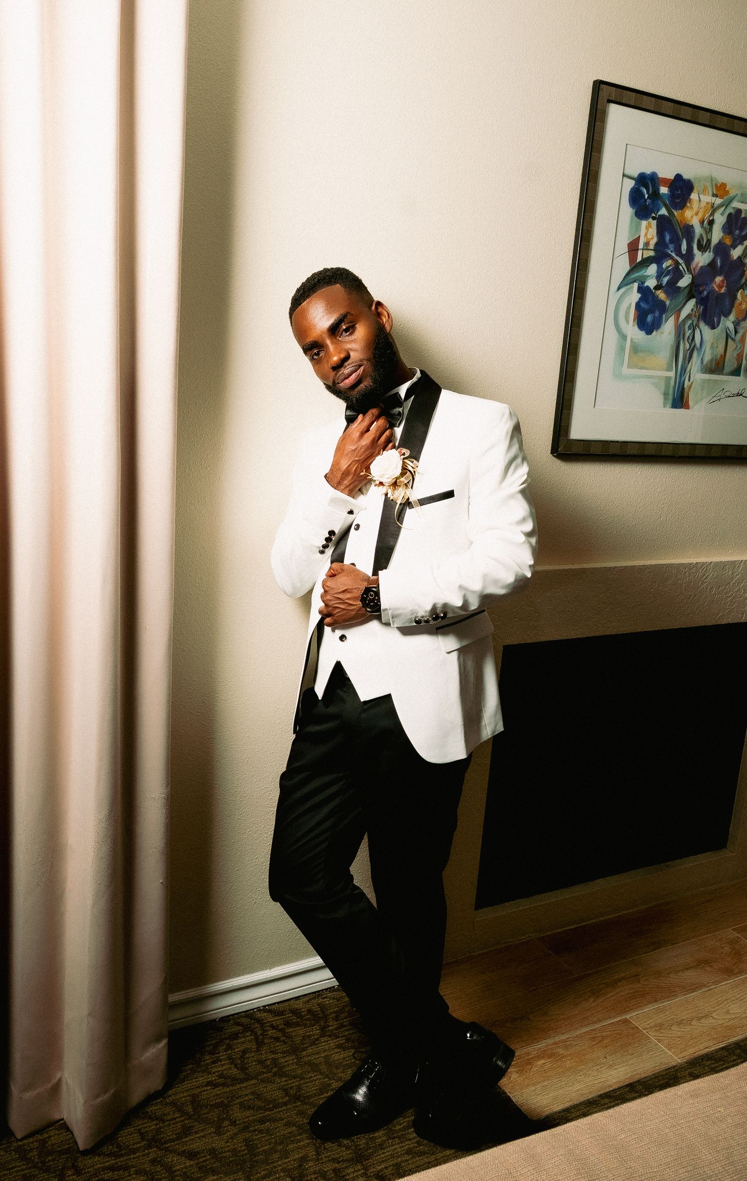 A person in a white tuxedo jacket and black pants poses dramatically against a wall in an interior hallway.
