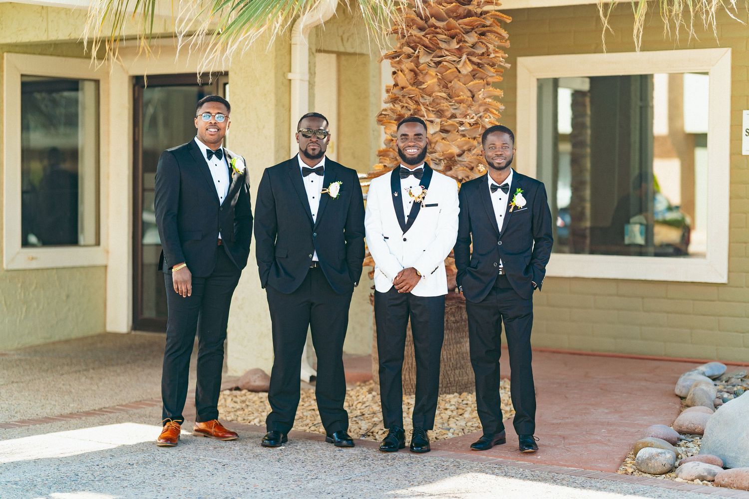 Four groomsmen in formal attire pose together outdoors in front of a beige building with palm trees.