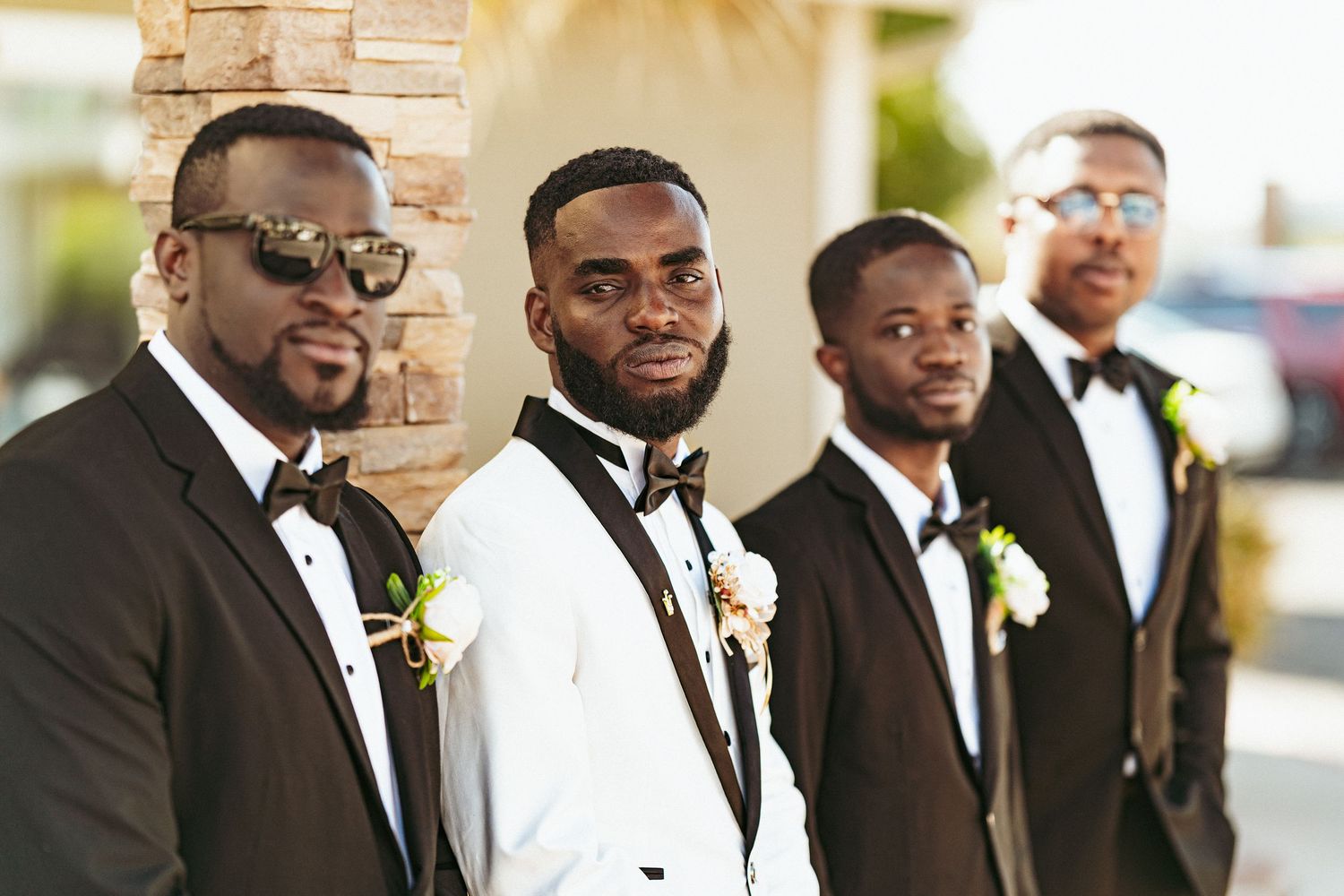 A wedding party in formal attire poses together outdoors with the groomsmen wearing black tuxedos.