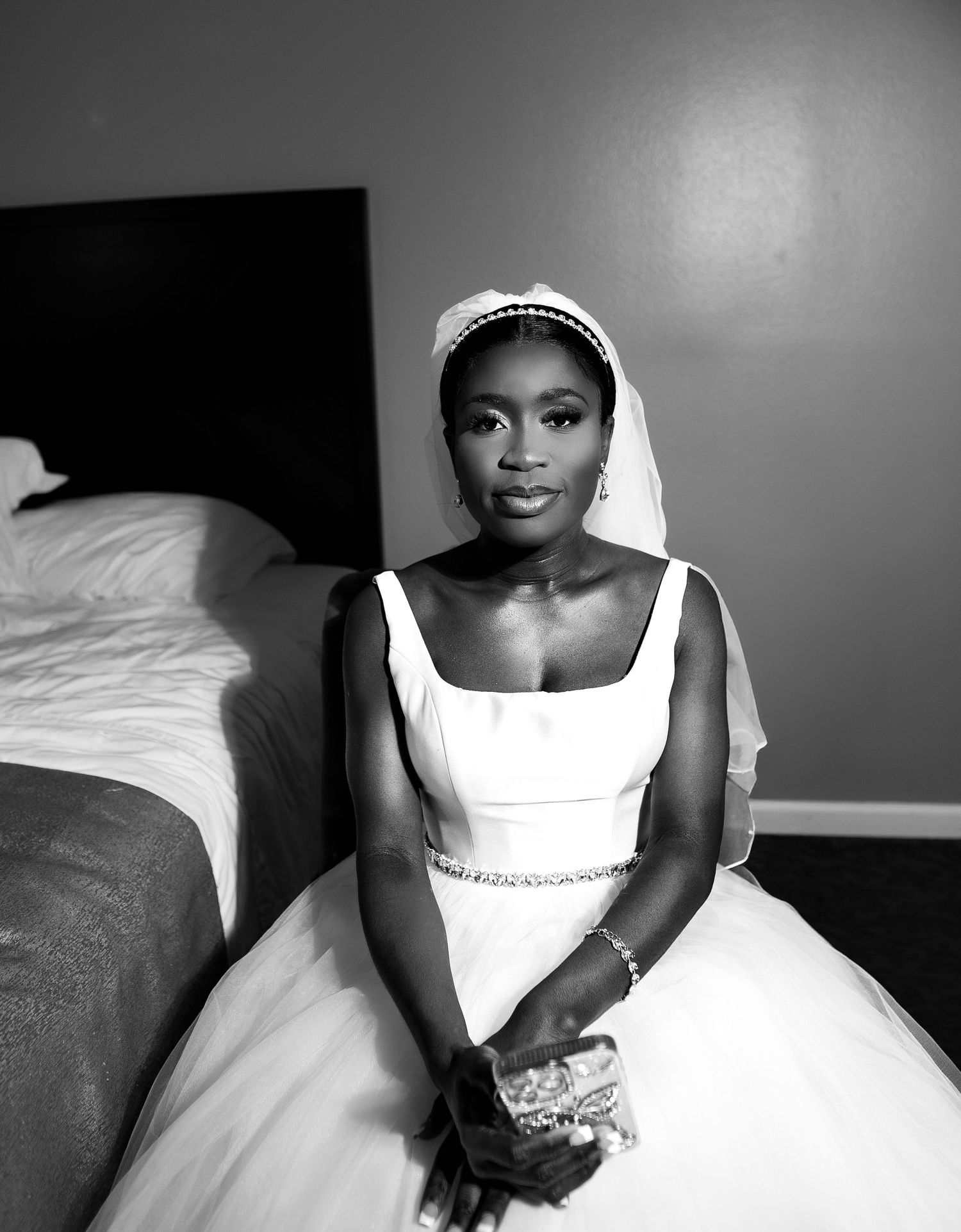 A black and white portrait of a bride in a classic white wedding dress with a veil, sitting on the edge of a bed.