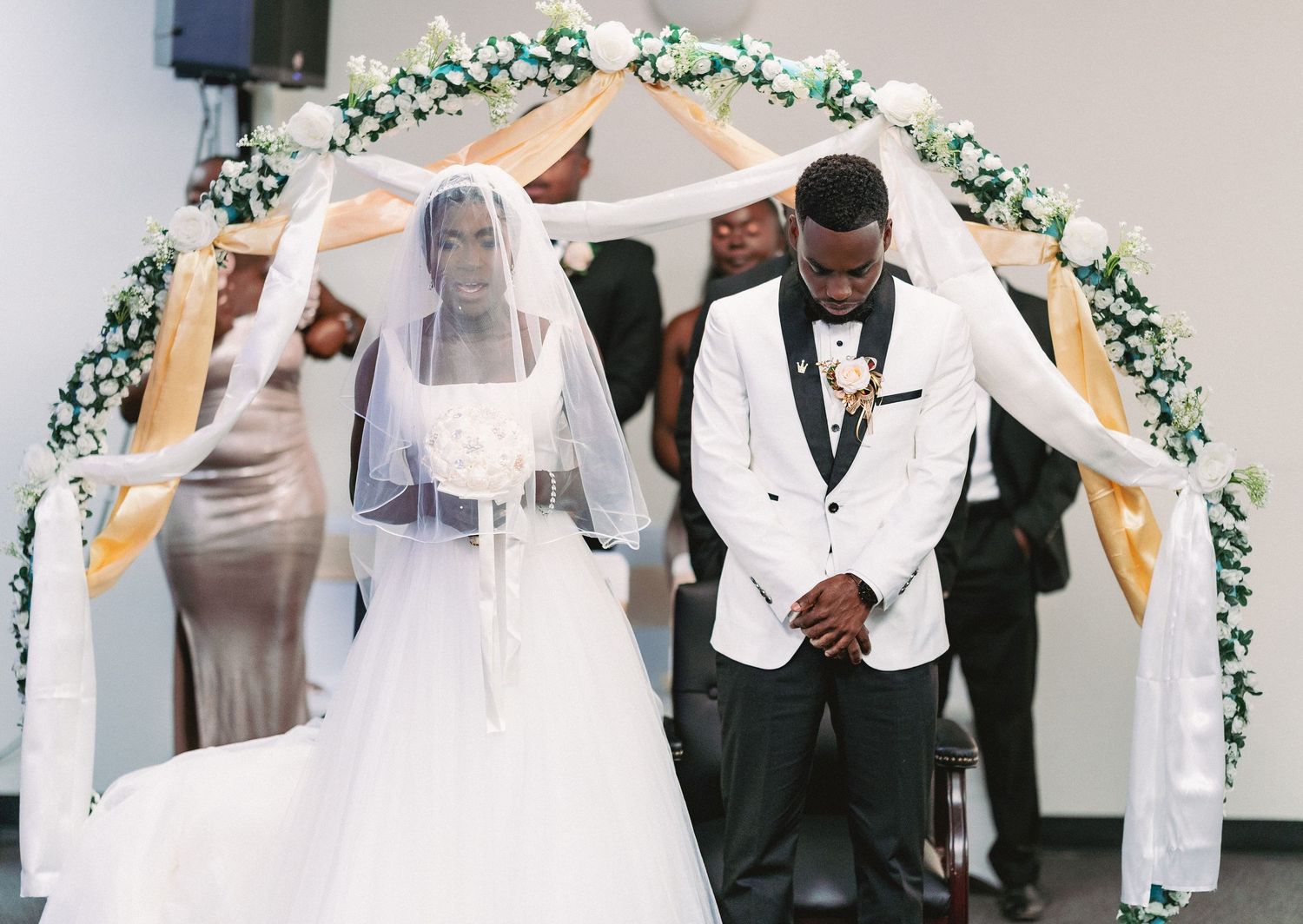 Wedding ceremony under a white floral arch with bridesmaids in light dresses and groomsmen in dark suits.