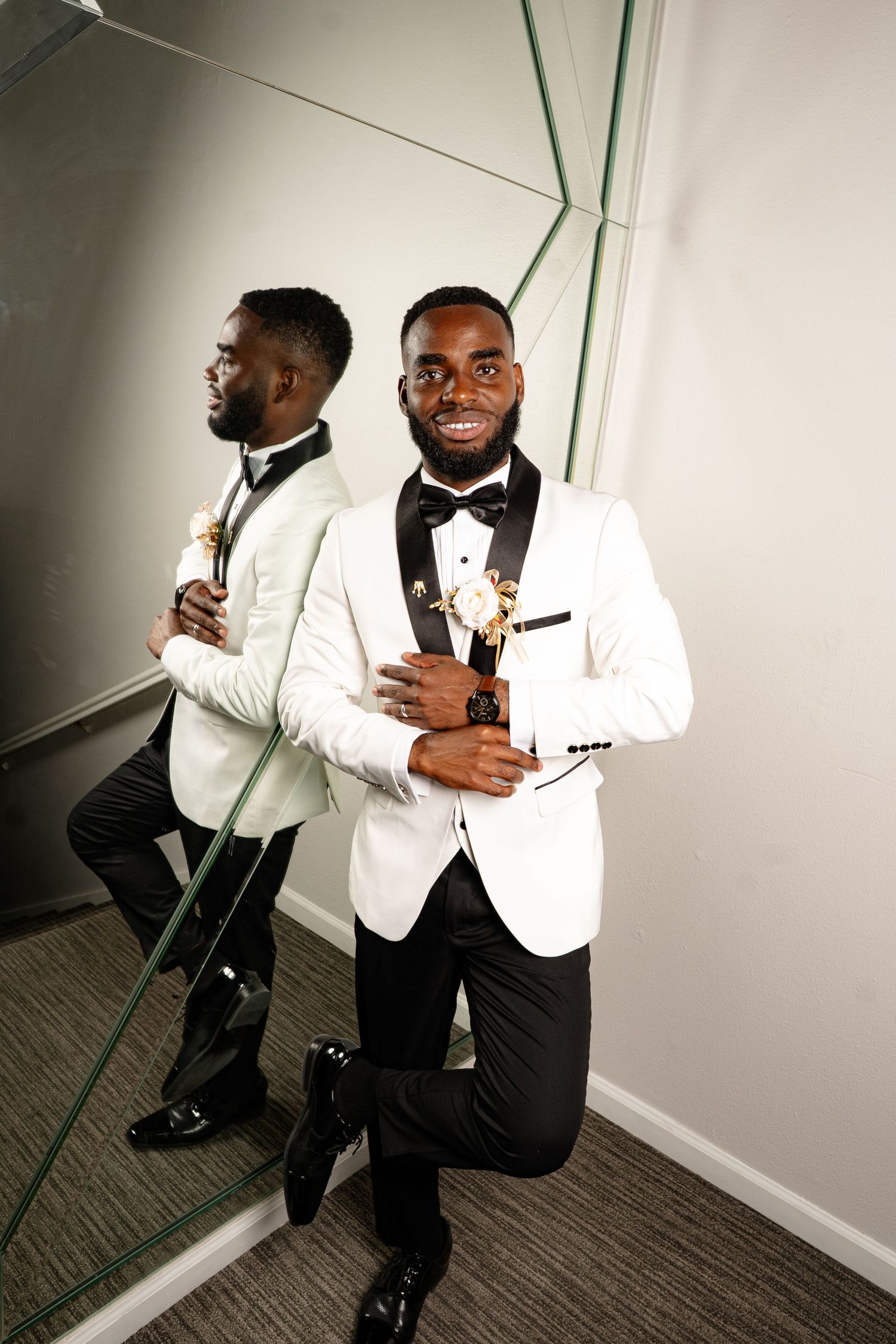 Groom in white tuxedo poses against mirror creating artistic reflections.