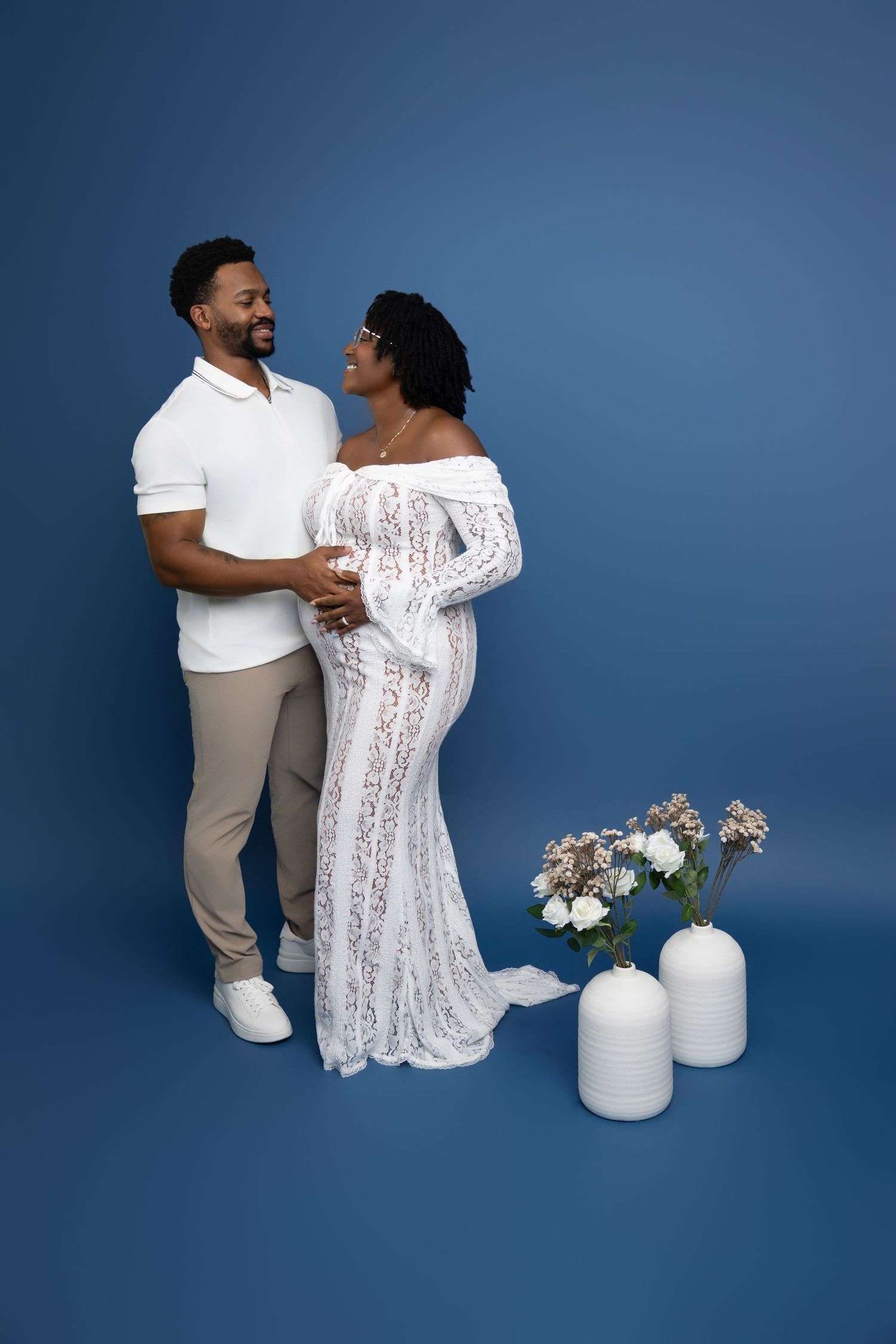 A couple in white and beige outfits poses against a blue backdrop with white pumpkins and floral decorations.