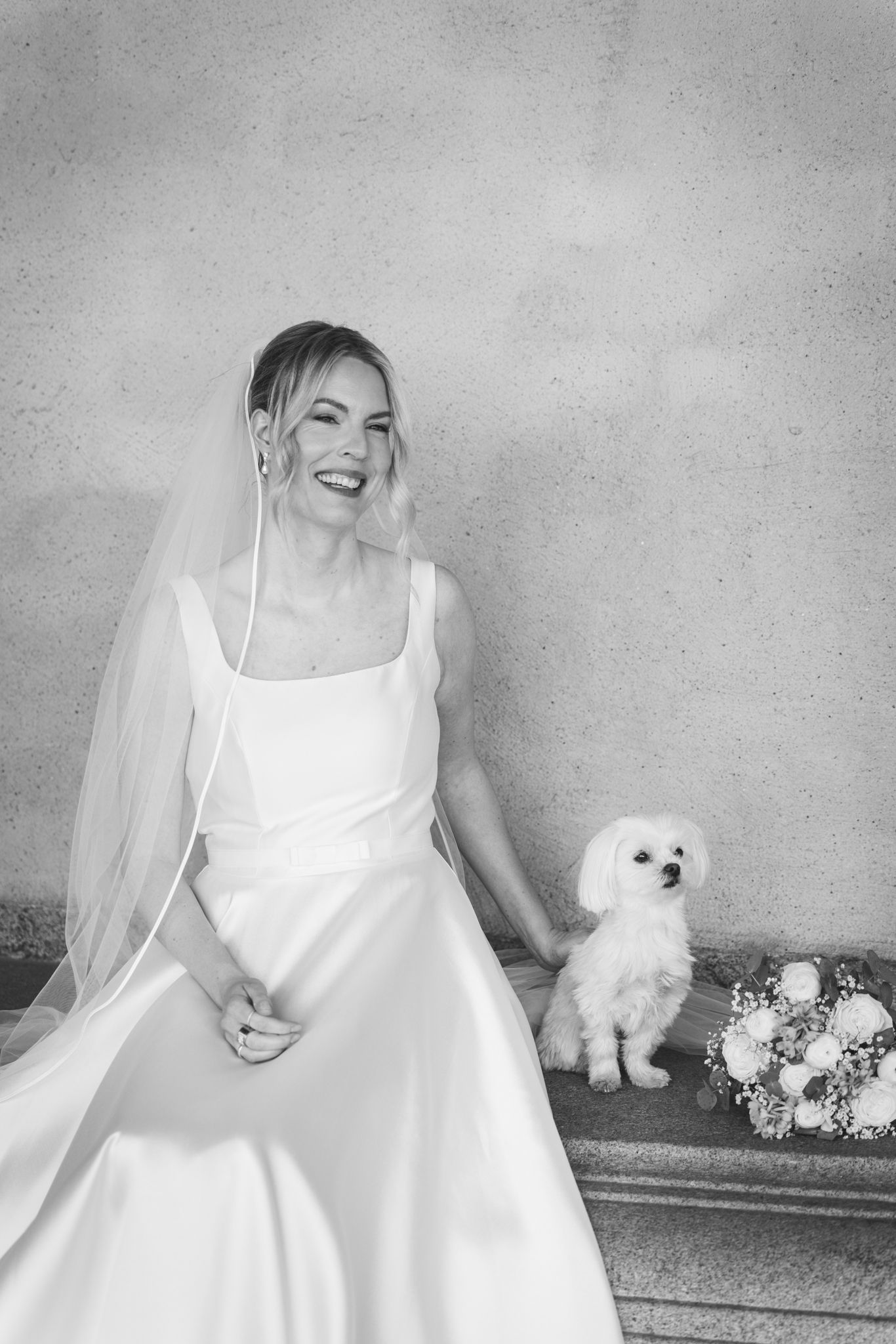 Black and white photo of a smiling bride in a white gown and veil sitting beside a small white dog and bouquet.