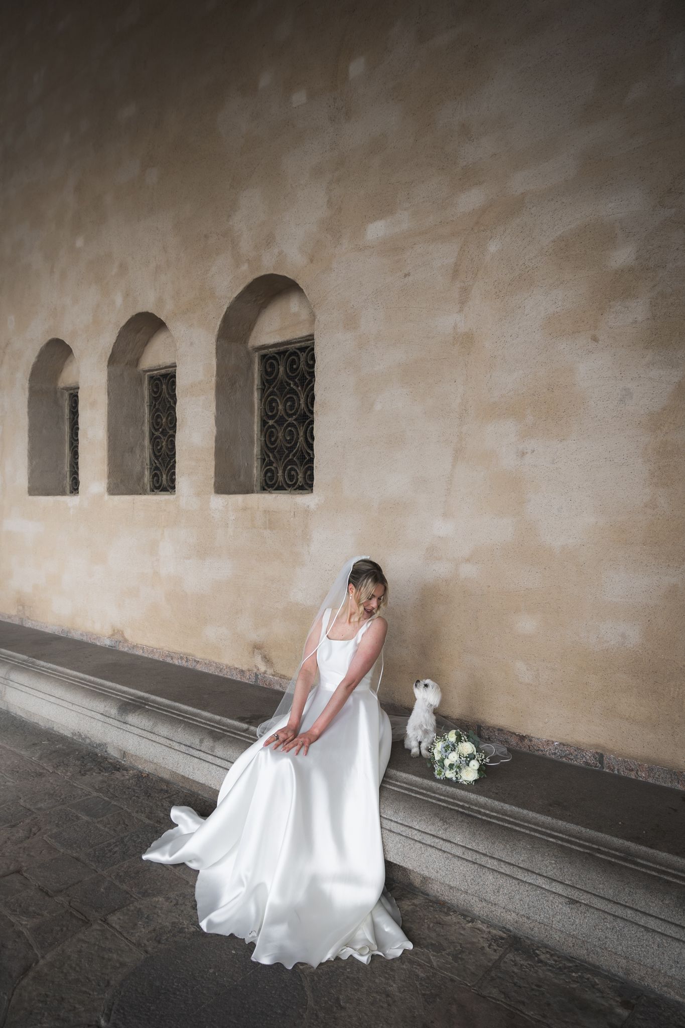Elegant bride in a flowing white wedding gown stands against a rustic stone wall with arched windows, holding a white bouquet.