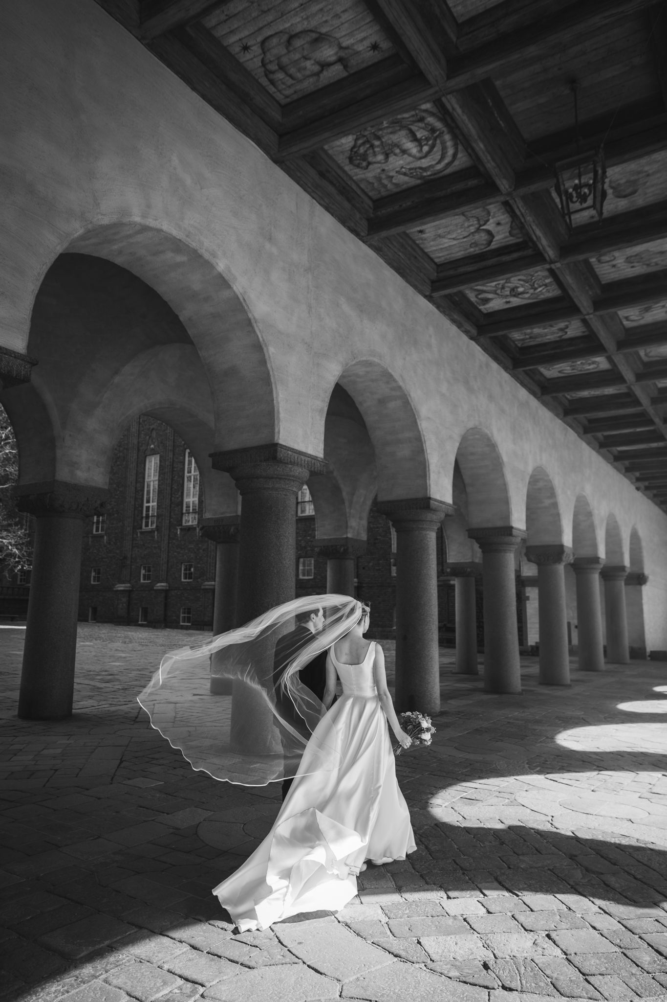 Black and white photo of a bride in a flowing gown with veil billowing under ornate arched colonnade.