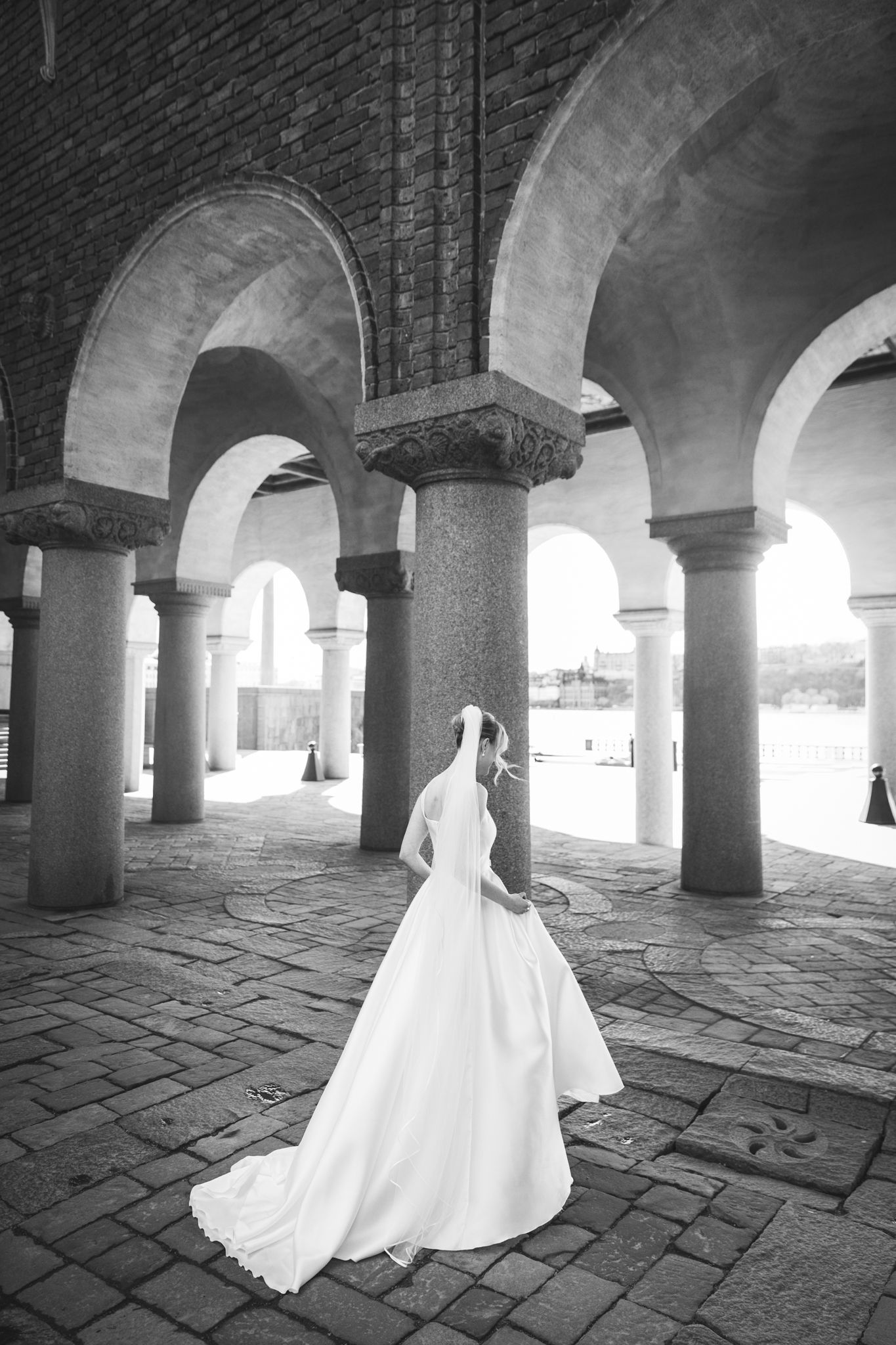 Black and white photo of a bride in a flowing white gown and veil standing among stone arches and columns.