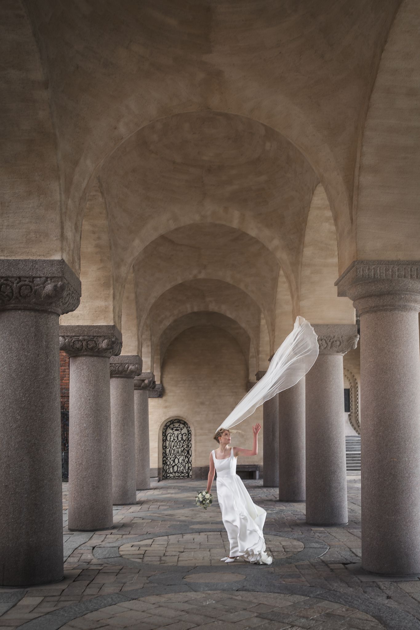 Bride in white gown with flowing veil caught by wind, standing among stone columns under arched ceiling.