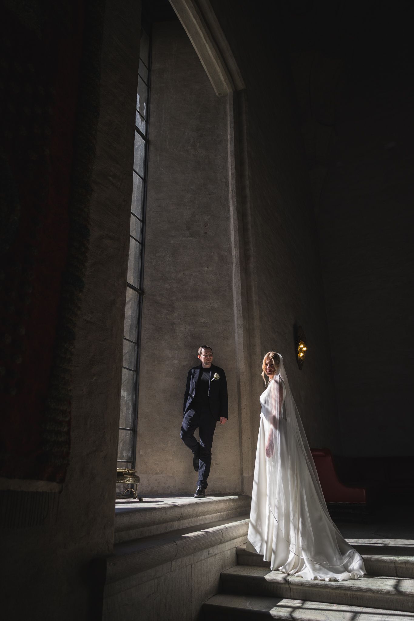 Bride and groom posing dramatically in a dark elegant venue, with light streaming through tall windows.