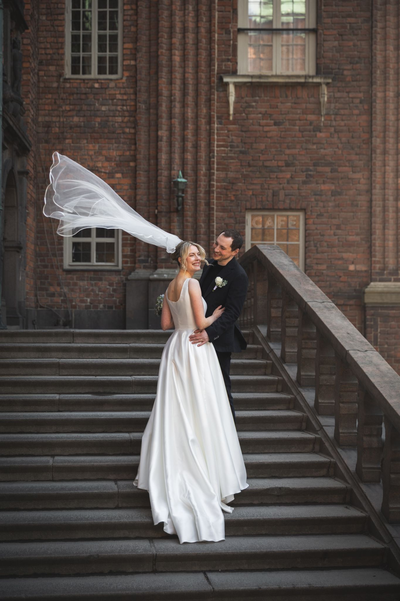 Bride in white gown with veil blowing in wind stands with groom on stone steps of brick building.