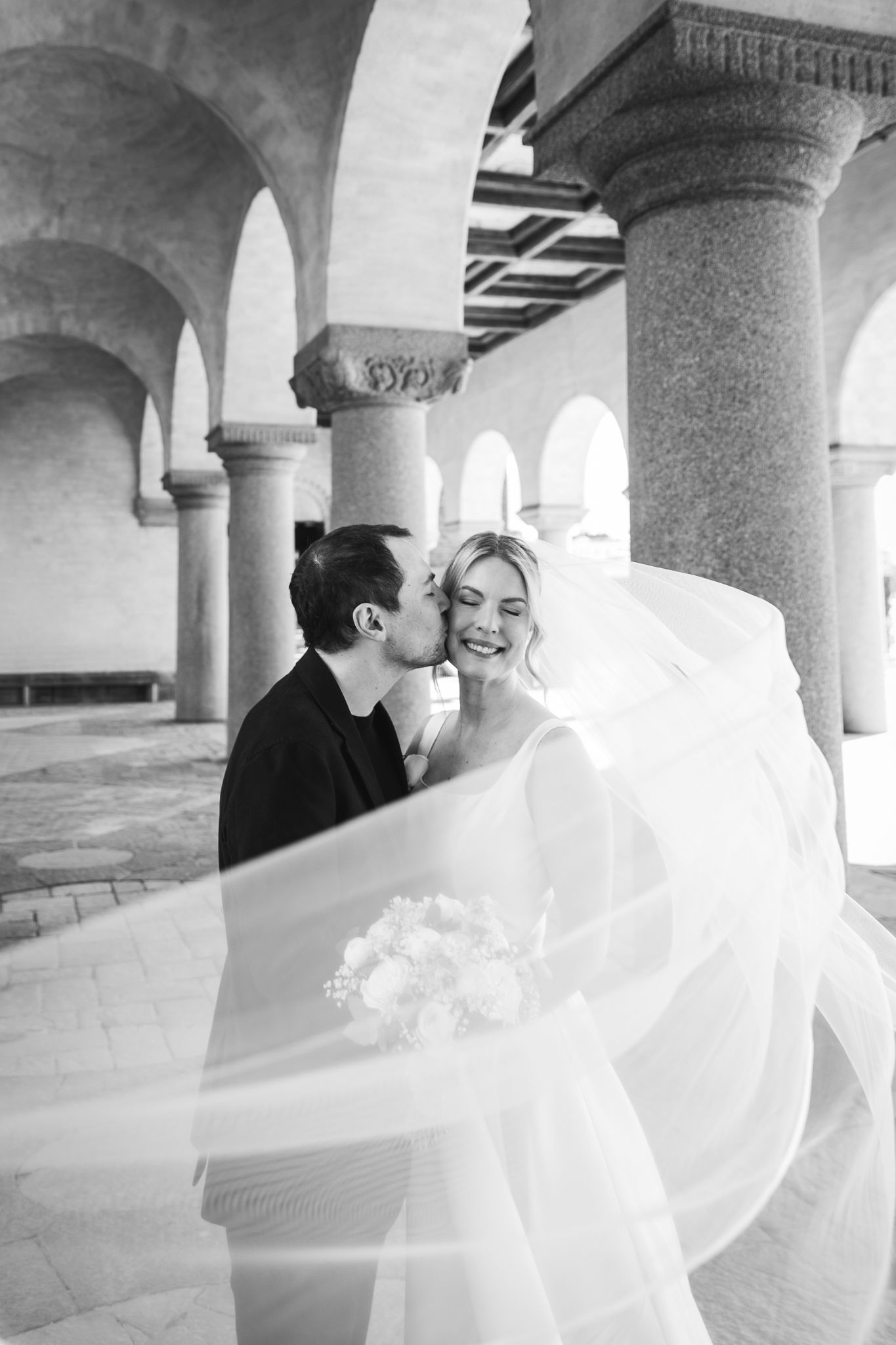Black and white wedding photo of bride and groom sharing a romantic moment under elegant stone arches, veil flowing in the wind.