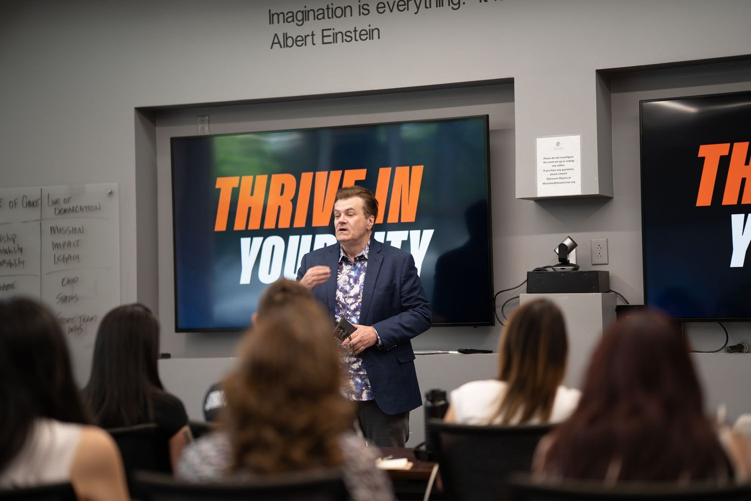 A speaker presents to an audience in a professional setting with digital displays showing "Thrive In" text in the background.