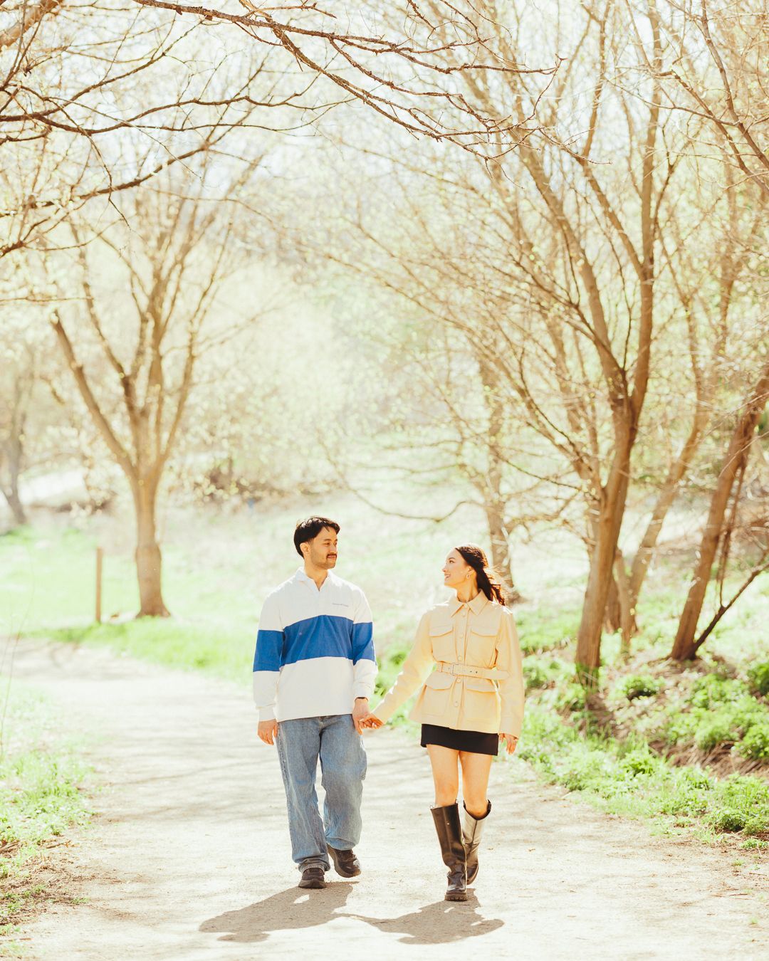 Couple walking hand in hand down a tree-lined path in soft sunlight.
