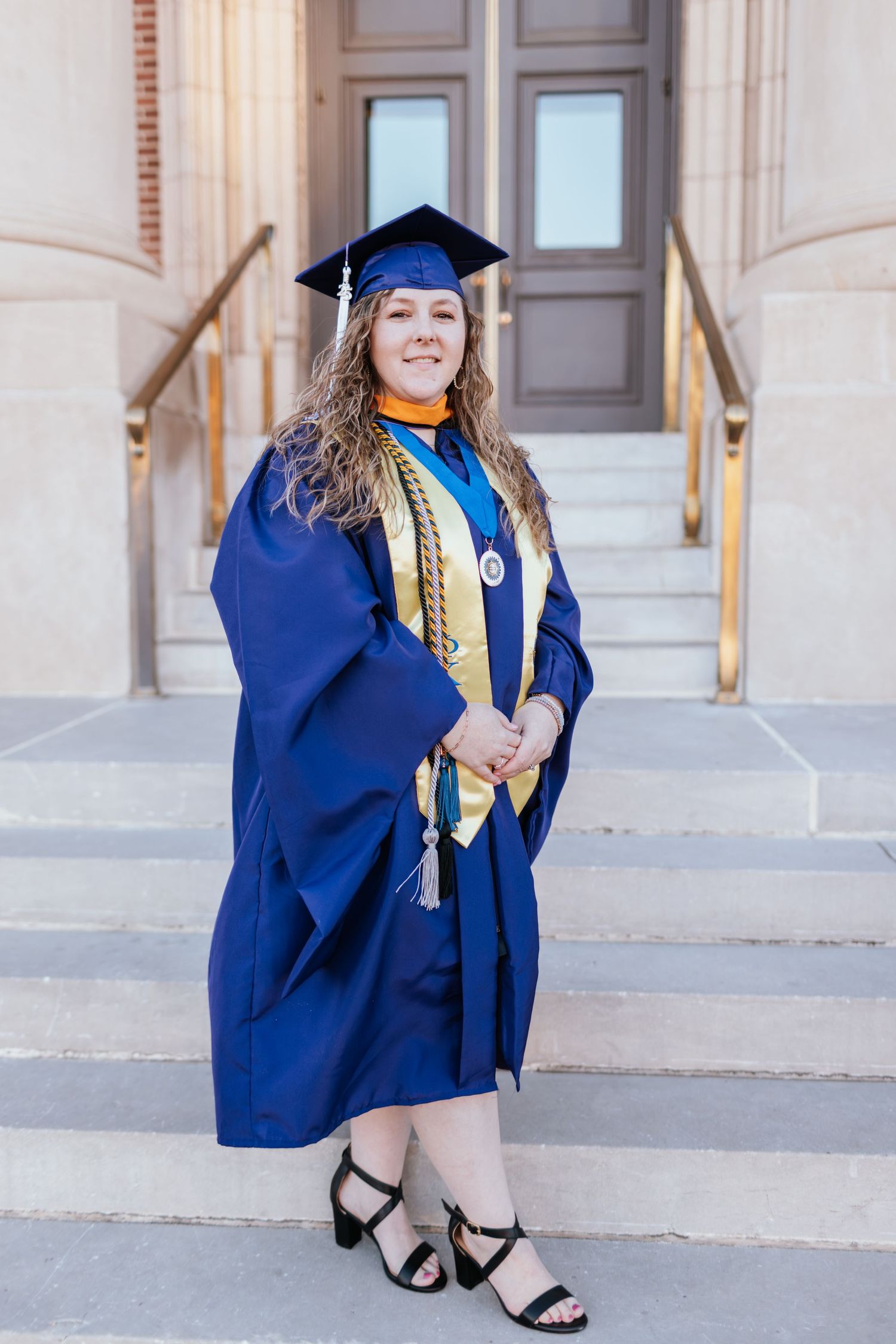 Graduate in blue cap and gown poses on stone steps with gold honor cords during graduation photo session.