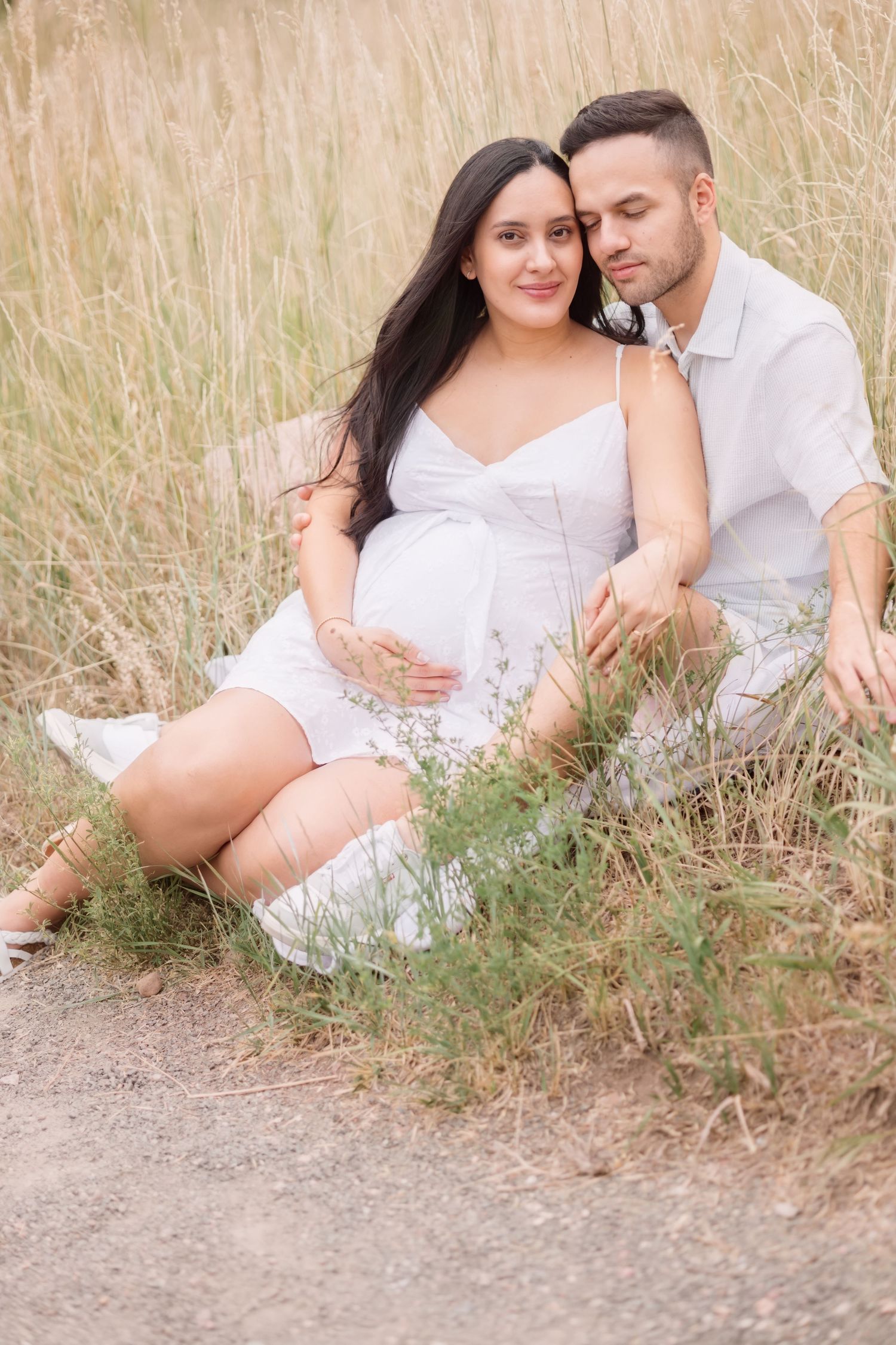 Couple in white outfits sharing tender moments while lying in tall grass during outdoor maternity photoshoot.