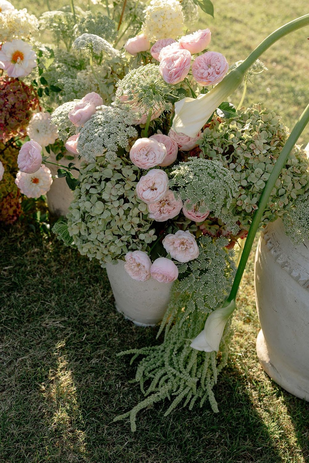 Delicate pink roses and white flowers arranged in ceramic pots with cascading greenery on a grassy lawn in soft sunlight.