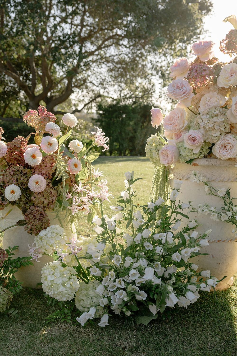 Lush outdoor wedding floral installation with pink roses, white blooms and greenery in natural sunlight.