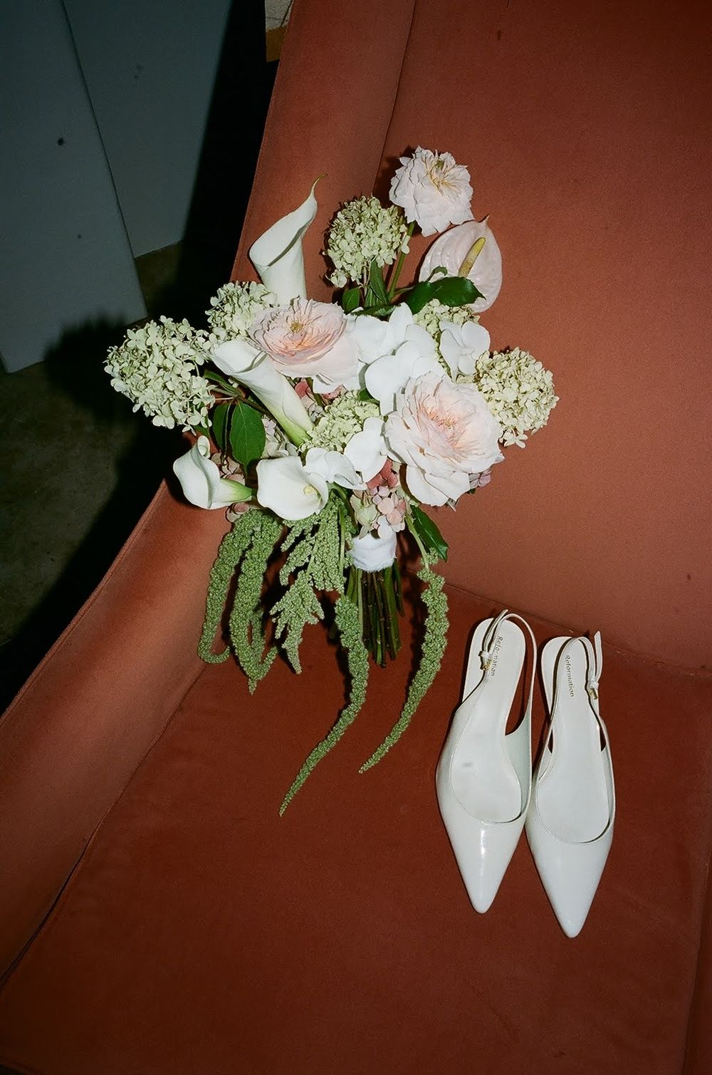 White bridal heels and delicate floral bouquet with pink and white flowers against a reddish-brown background.