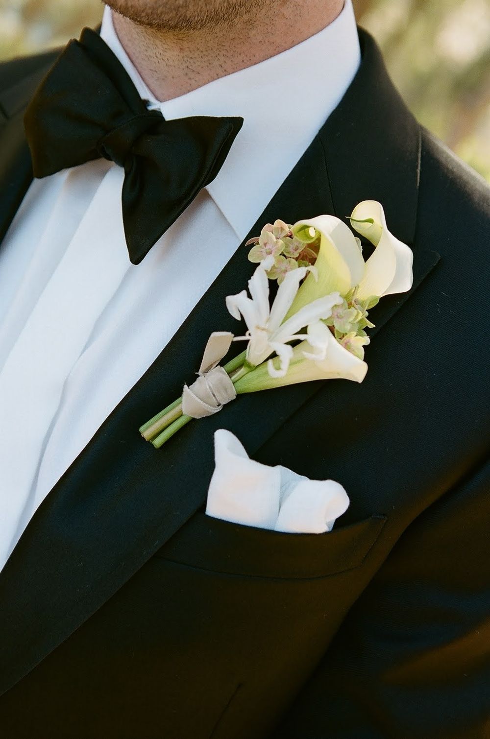 Close-up of classic black tuxedo with white boutonniere featuring calla lilies and a black bow tie for formal wedding attire.