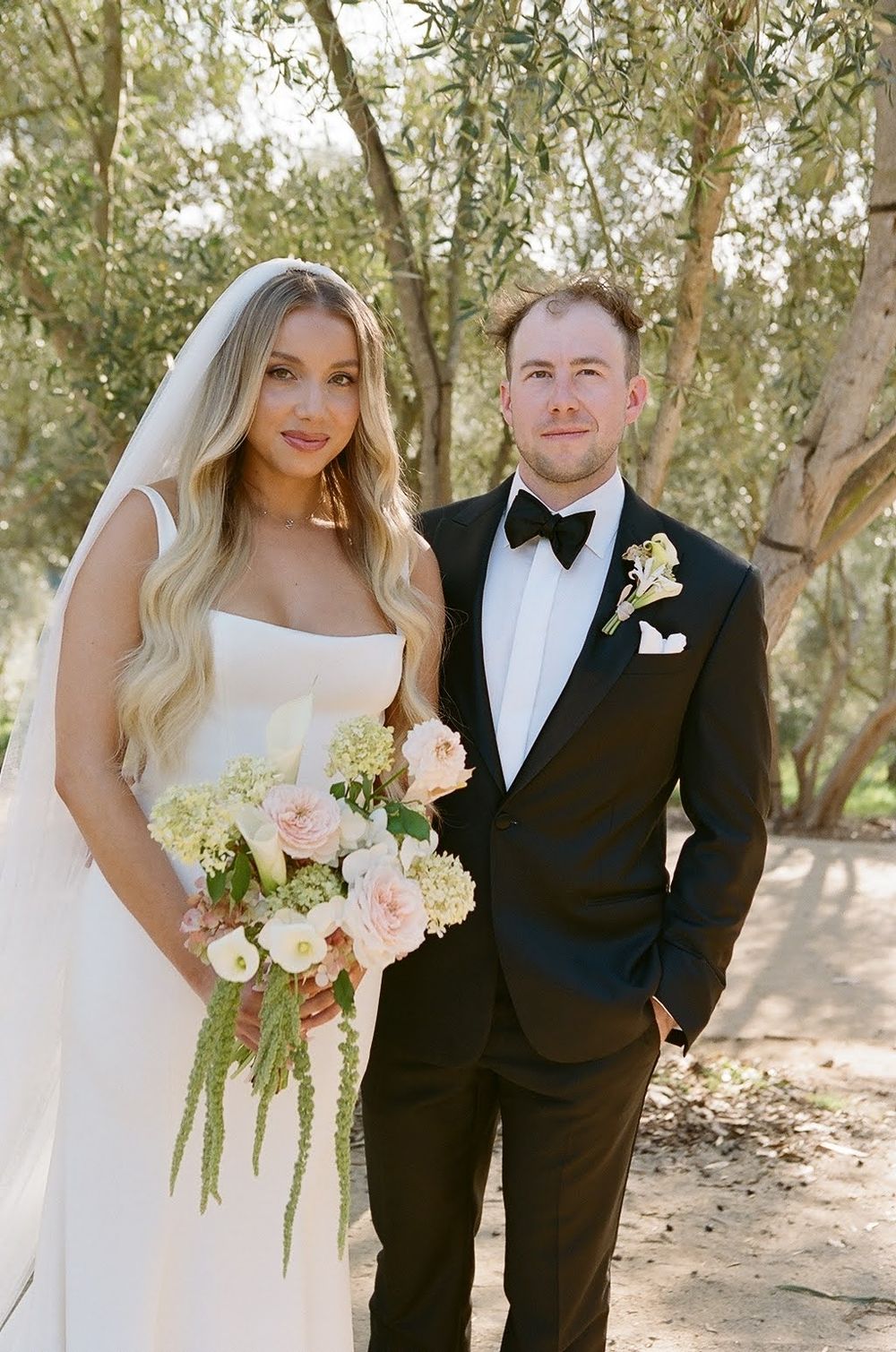 Newlywed couple poses outdoors among olive trees in formal black tuxedo and white wedding gown with blush bouquet.