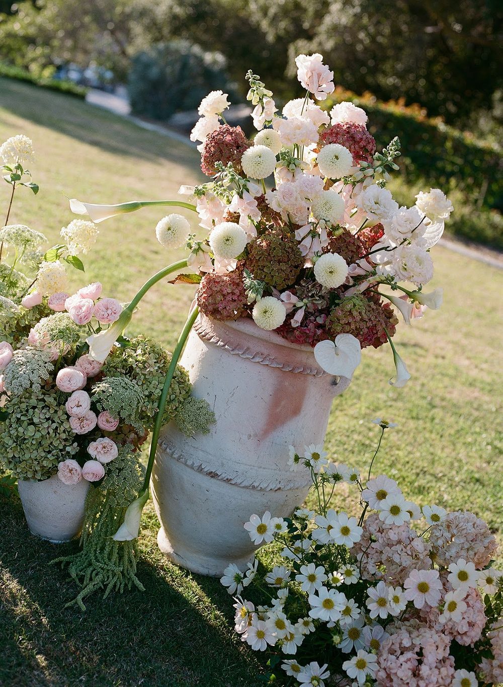 Rustic flower arrangements in ceramic vases featuring daisies, roses and wildflowers set on grass.