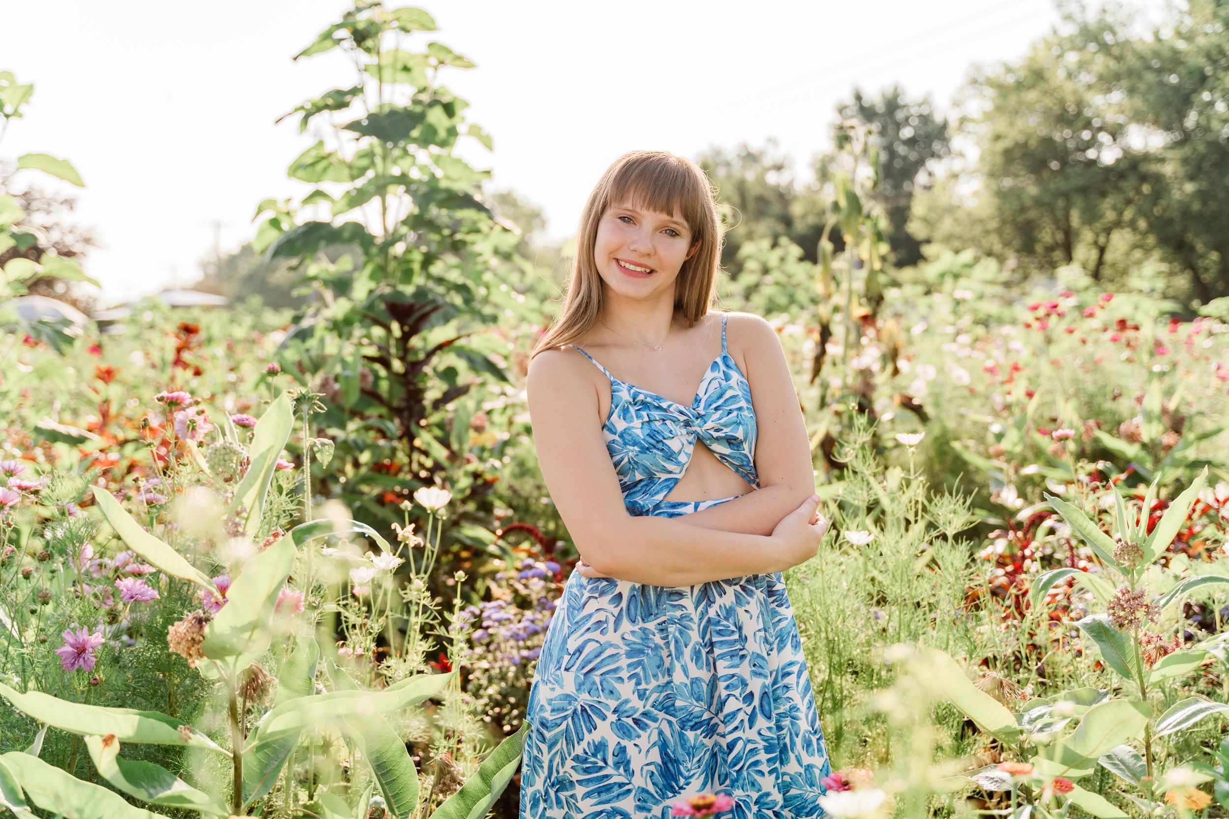 A person in a blue floral dress poses among colorful wildflowers and greenery in a sunny garden setting.