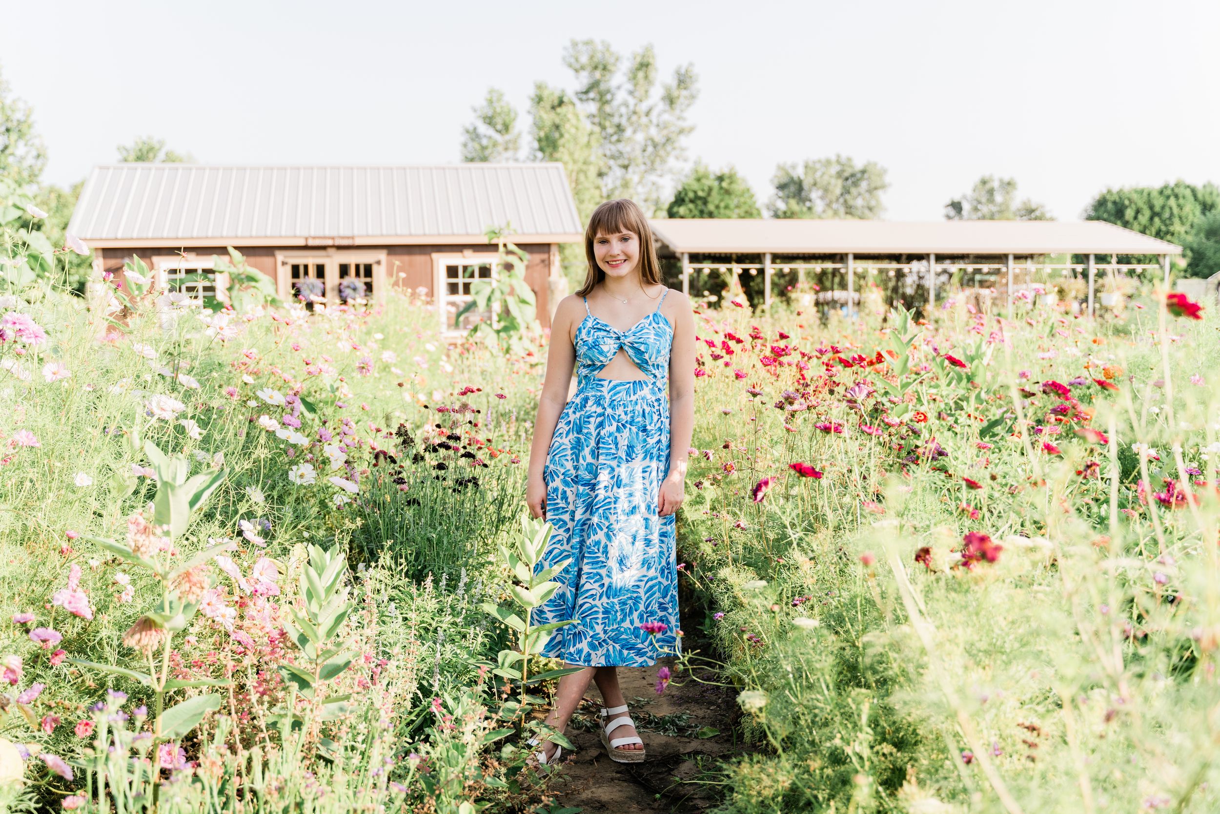 Someone in a blue floral sundress stands in a vibrant garden with red poppies and rustic buildings in the background.