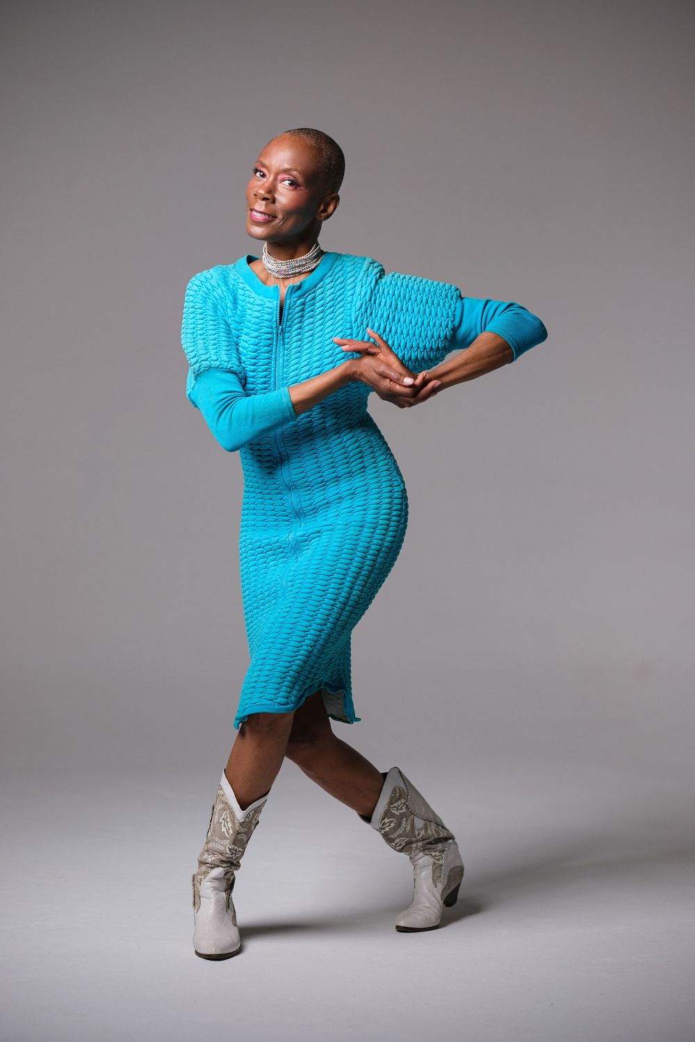 Energetic dance pose in bright blue sweater dress and white boots against grey background.