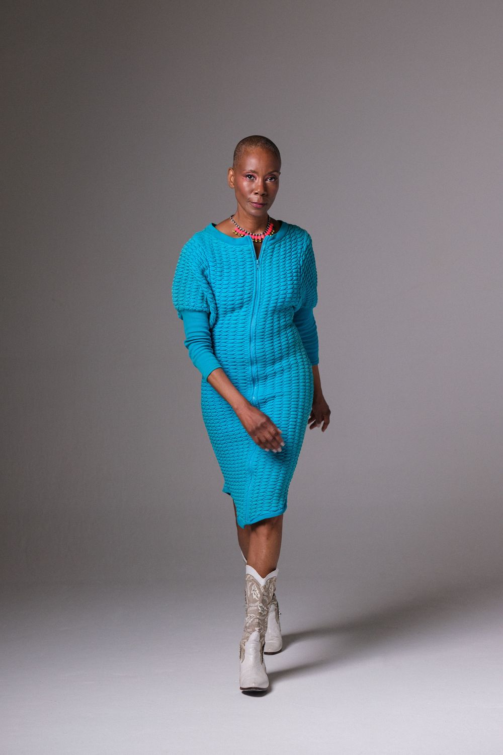Model in bright turquoise knit dress and white boots poses confidently against grey backdrop.
