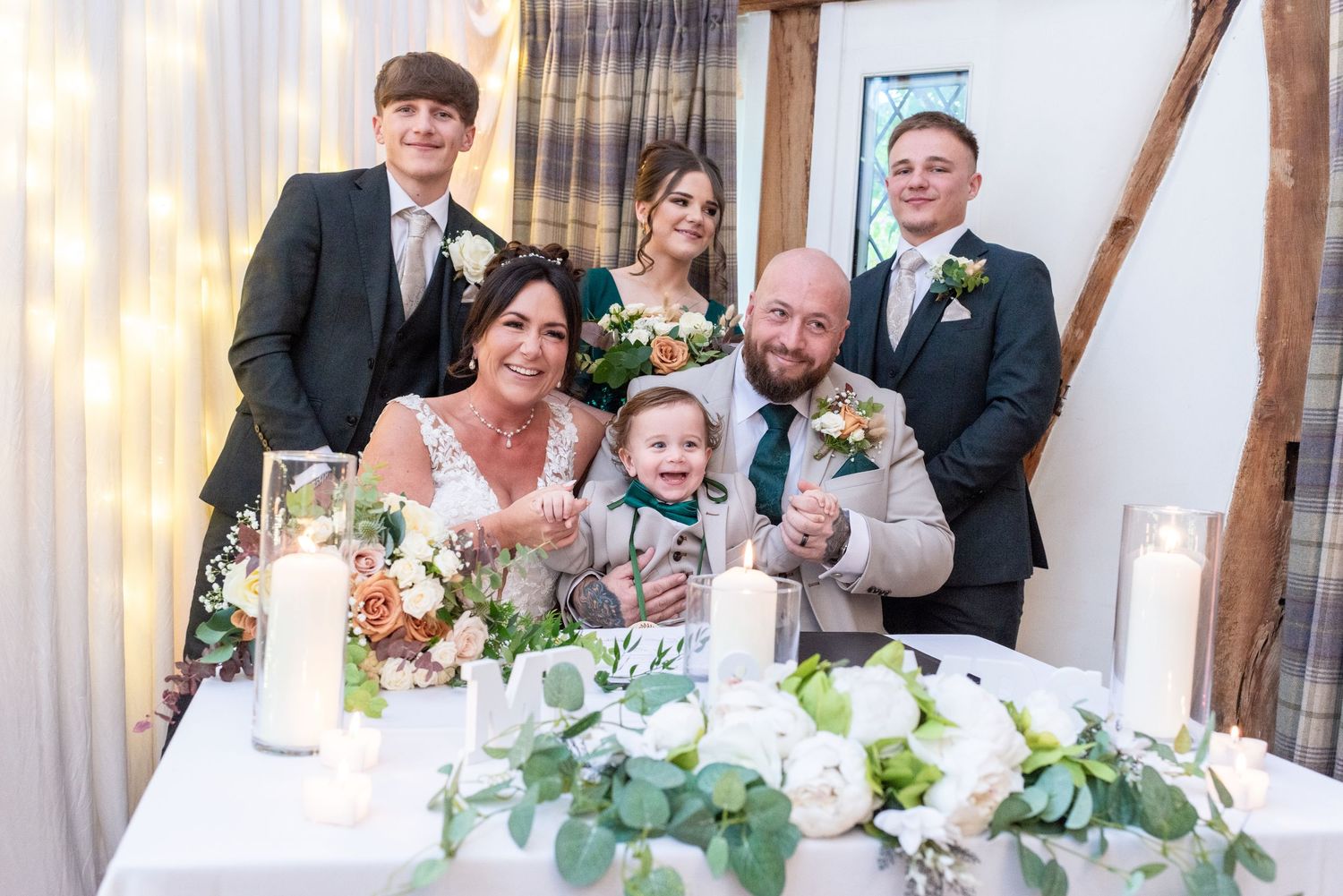 A joyful wedding party poses together at a decorated reception table with candles and floral centerpieces.