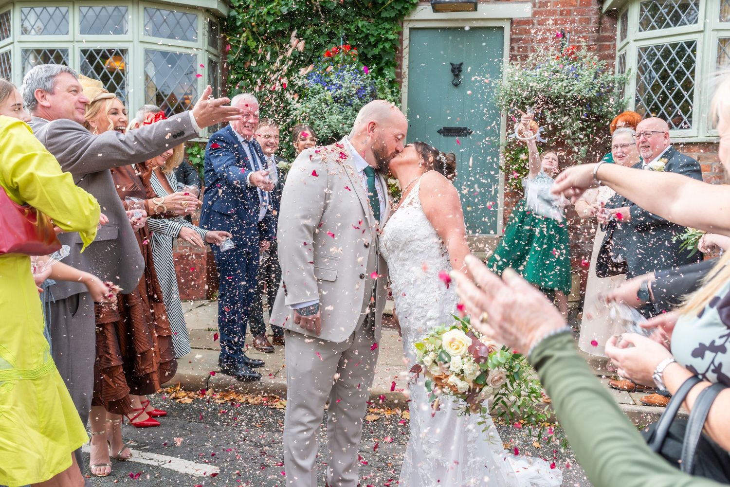 Wedding party showered with confetti outside a brick building with climbing vines and white trim.
