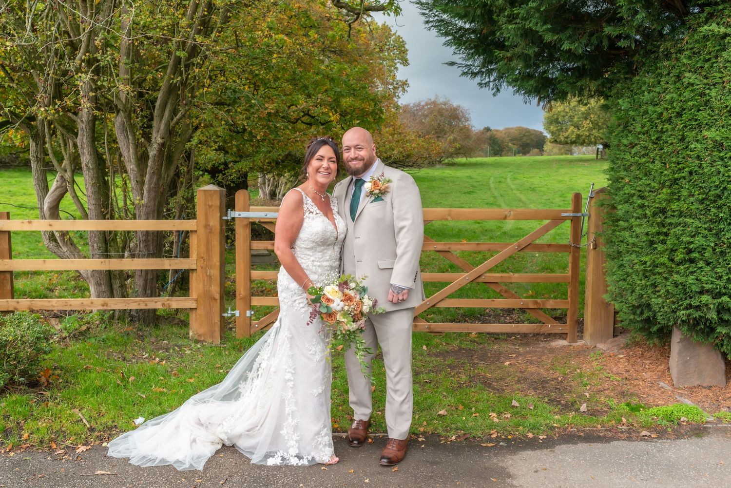 Wedding couple poses by rustic wooden fence in outdoor garden setting with autumn foliage.