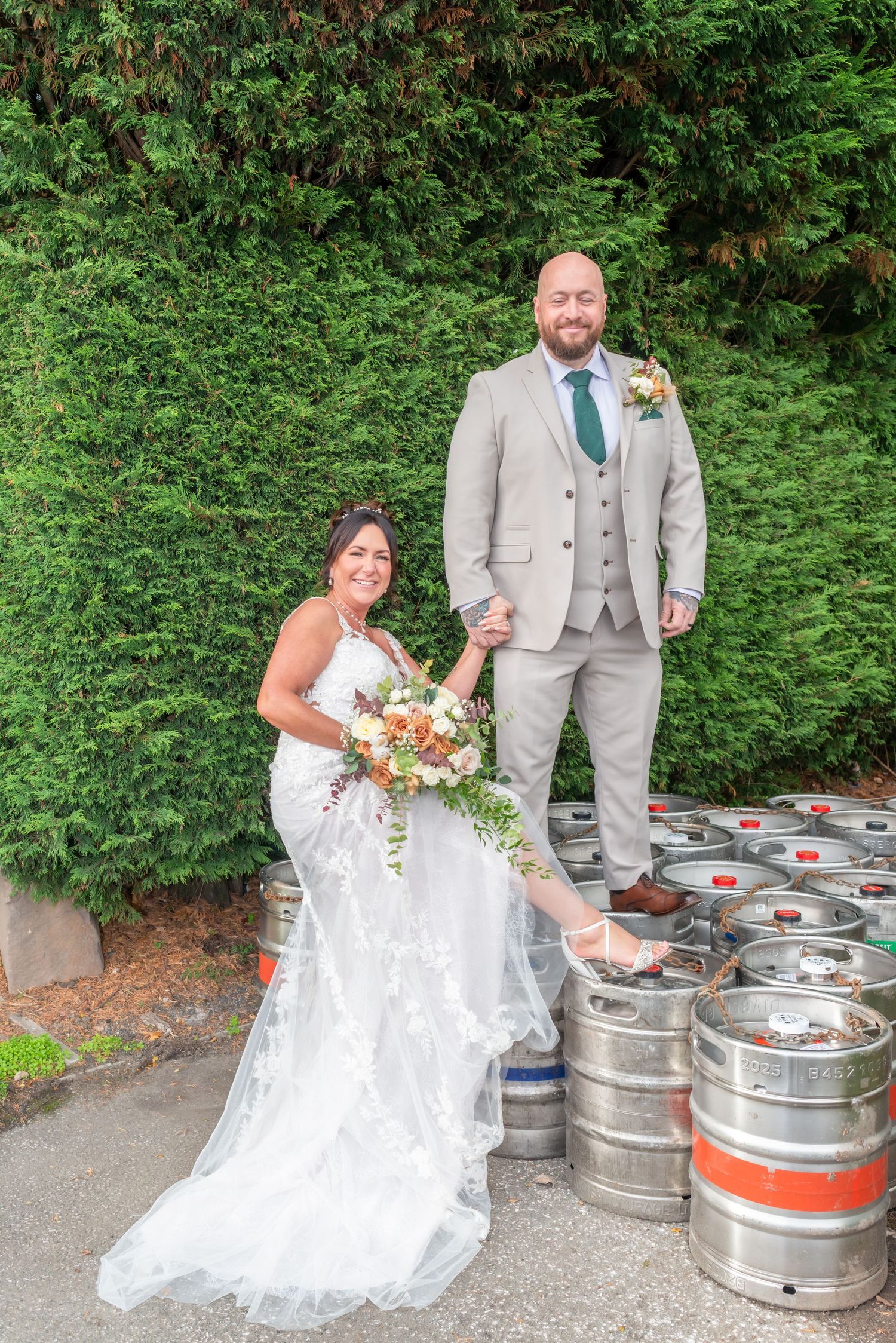 Wedding photos by beer kegs showing couple in grey suit and white dress with floral bouquet.