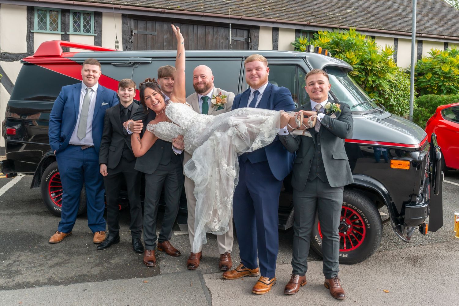 Wedding party celebrates with fun poses by parked cars outside brick building.