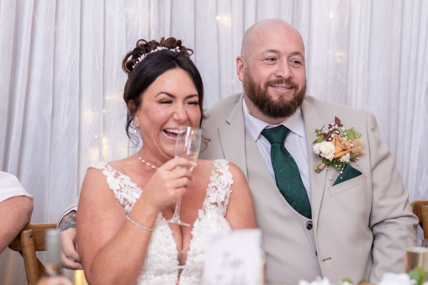 Wedding reception guests laughing and celebrating at tables with white decor and ambient lighting.