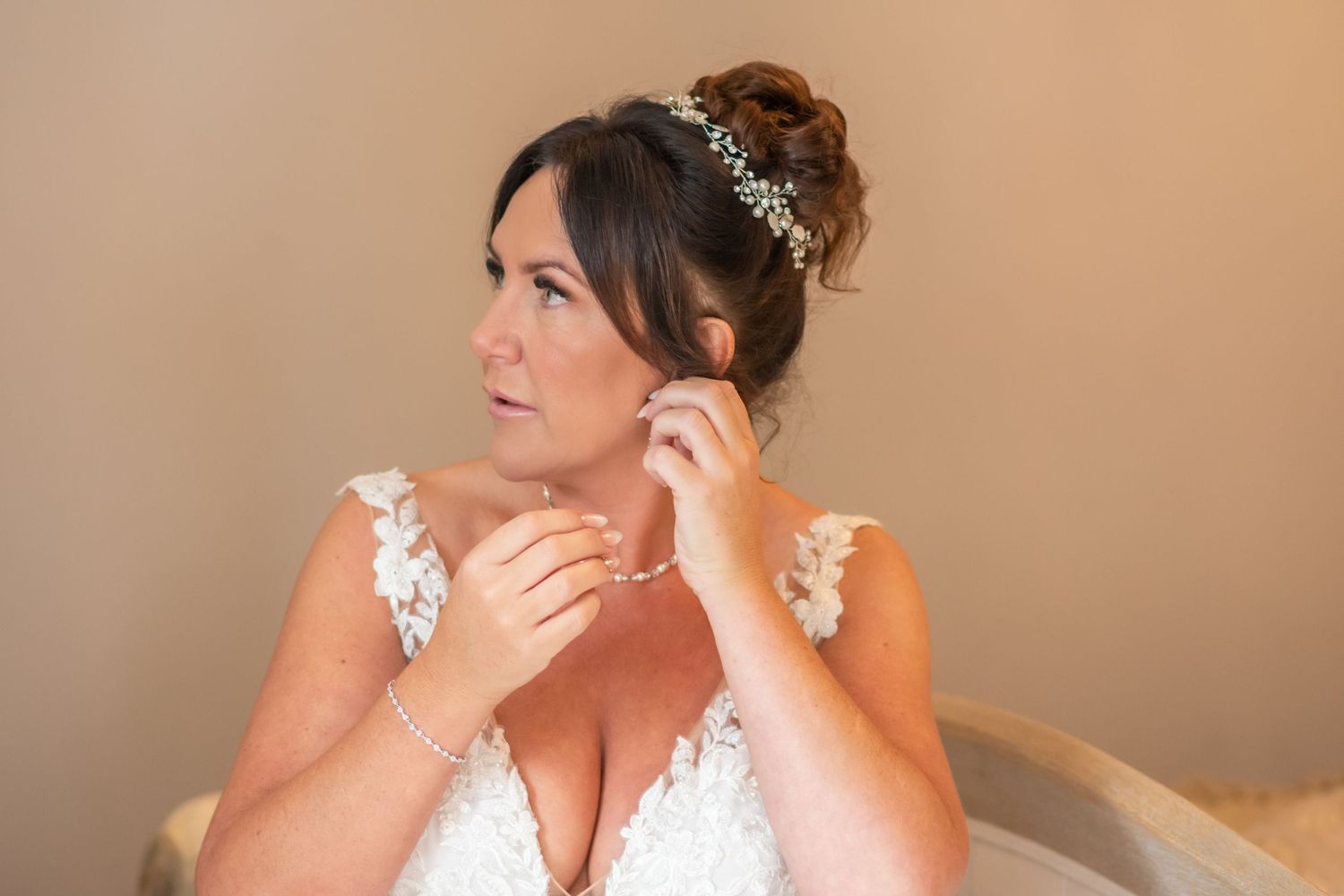 Bride in white lace wedding dress puts on delicate earrings while getting ready against neutral backdrop at the Plough Inn Eaton