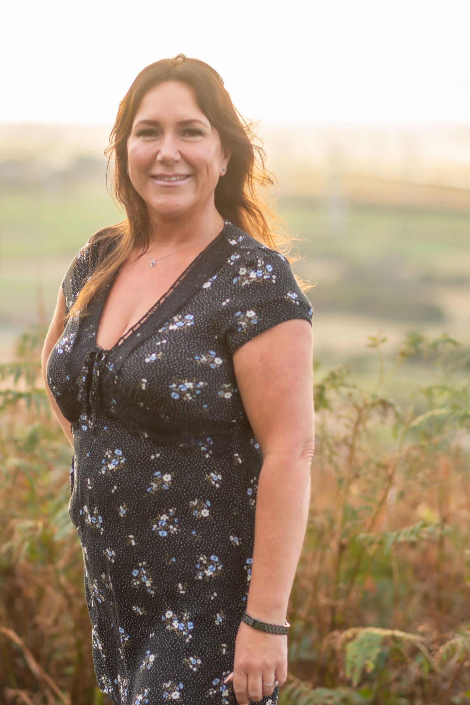 A person in a black floral dress poses outdoors in a field during golden hour.