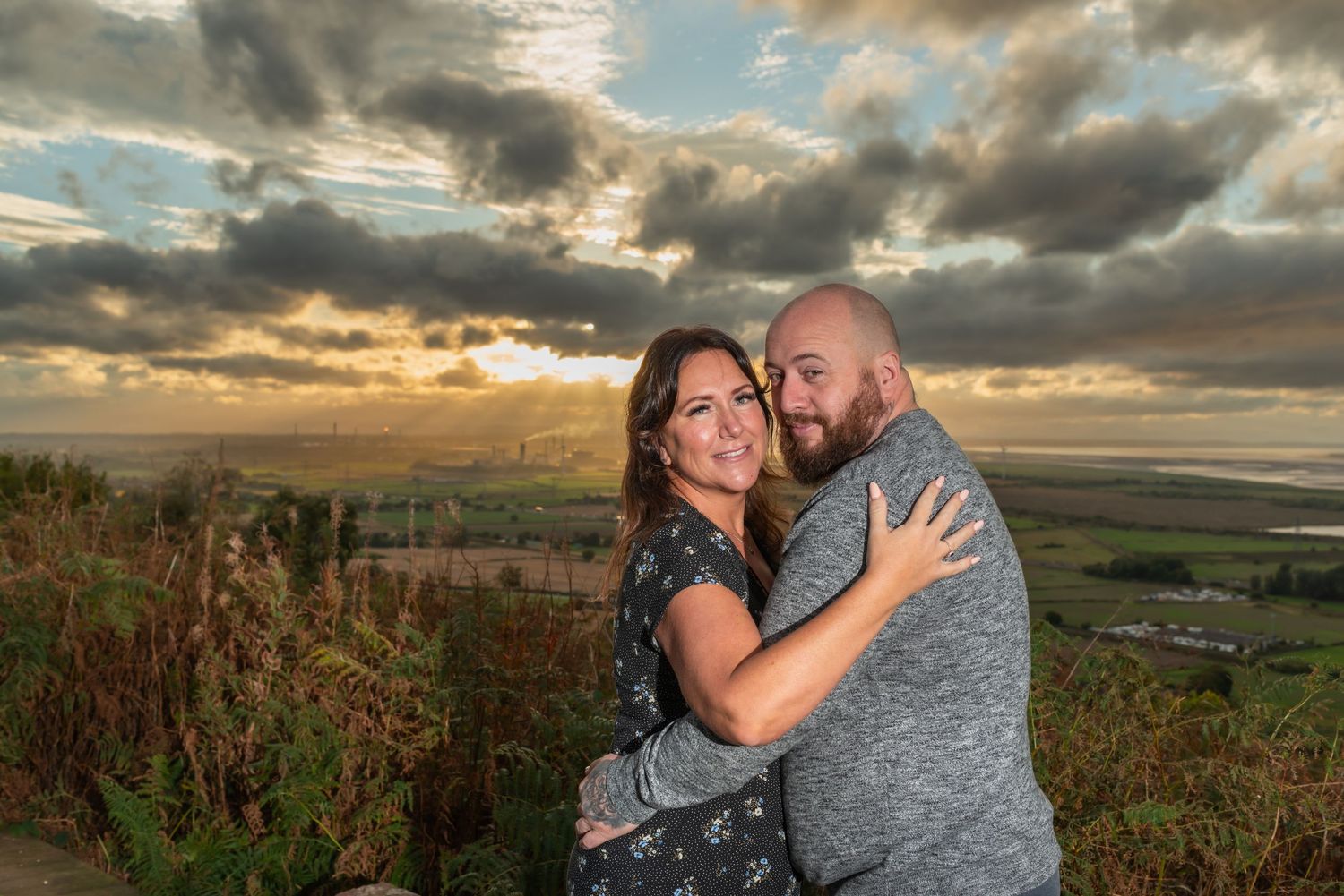 A couple embraces during a sunset photoshoot overlooking rolling hills and dramatic cloudy skies.