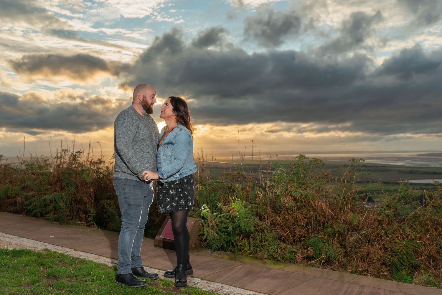 A couple shares a romantic moment at sunset on a hilltop overlooking a scenic countryside landscape.