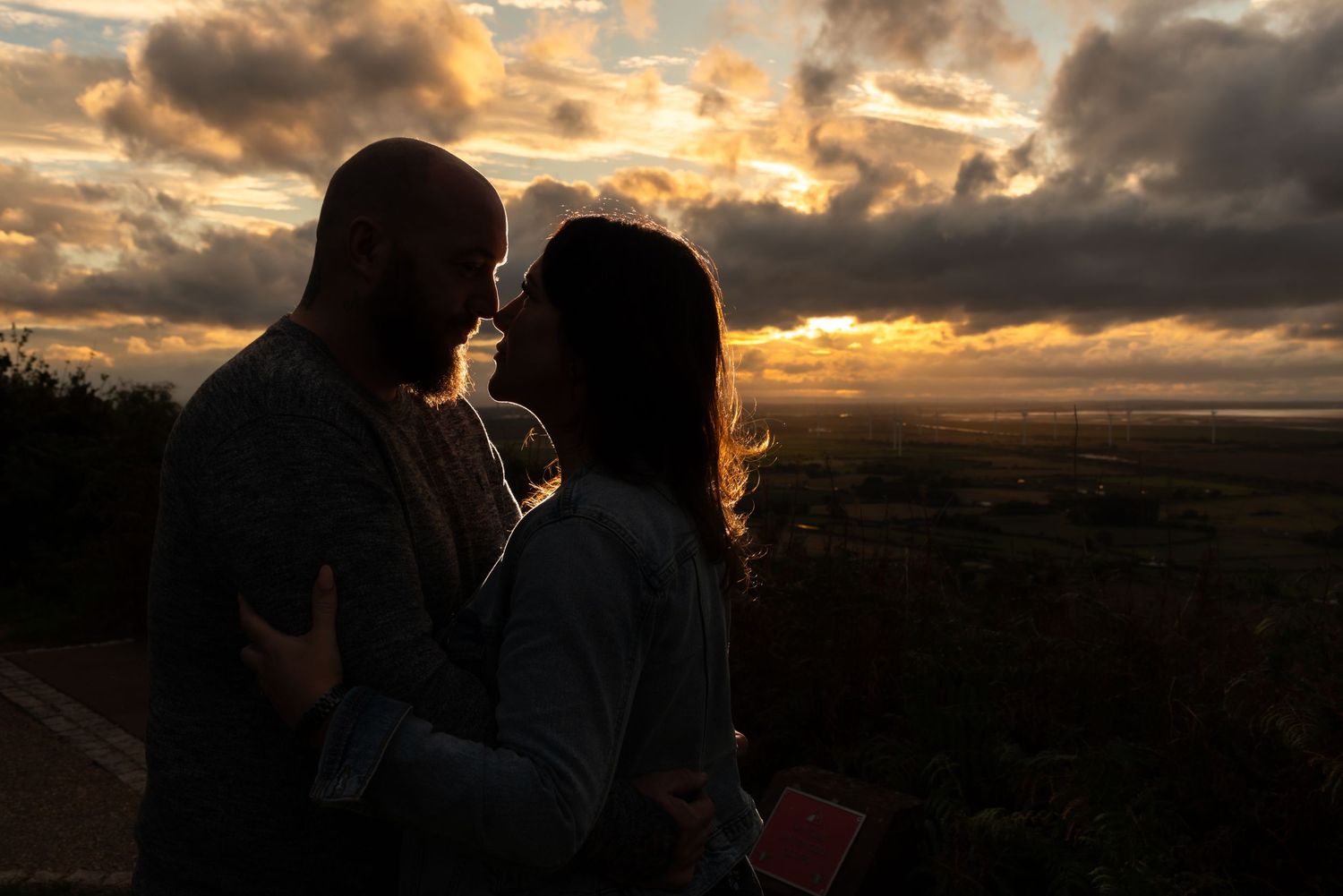 Two silhouettes embrace against a dramatic sunset sky with clouds and rolling hills in the background.