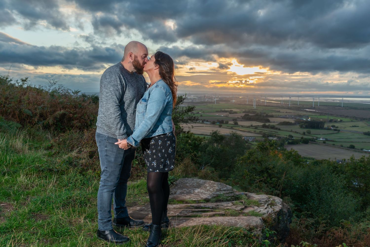 A couple embraces during sunset overlooking a scenic countryside valley with dramatic cloudy skies.