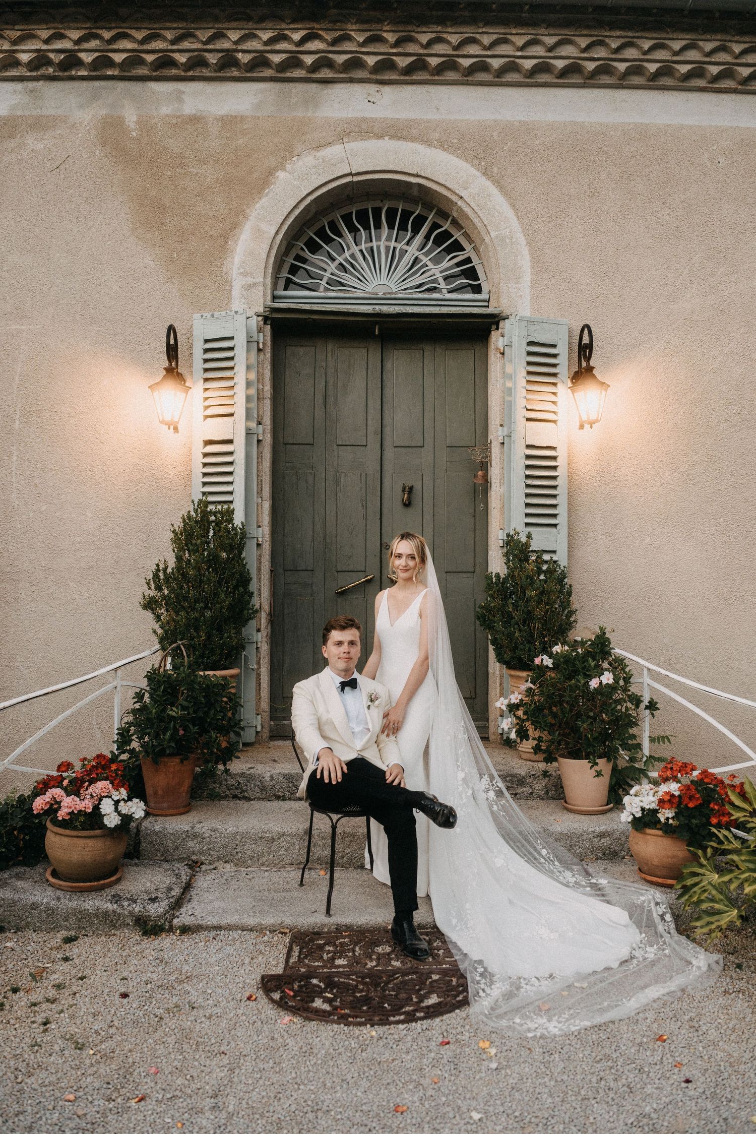 A wedding couple shares a lighthearted moment together on chairs outside a rustic building with shutters.