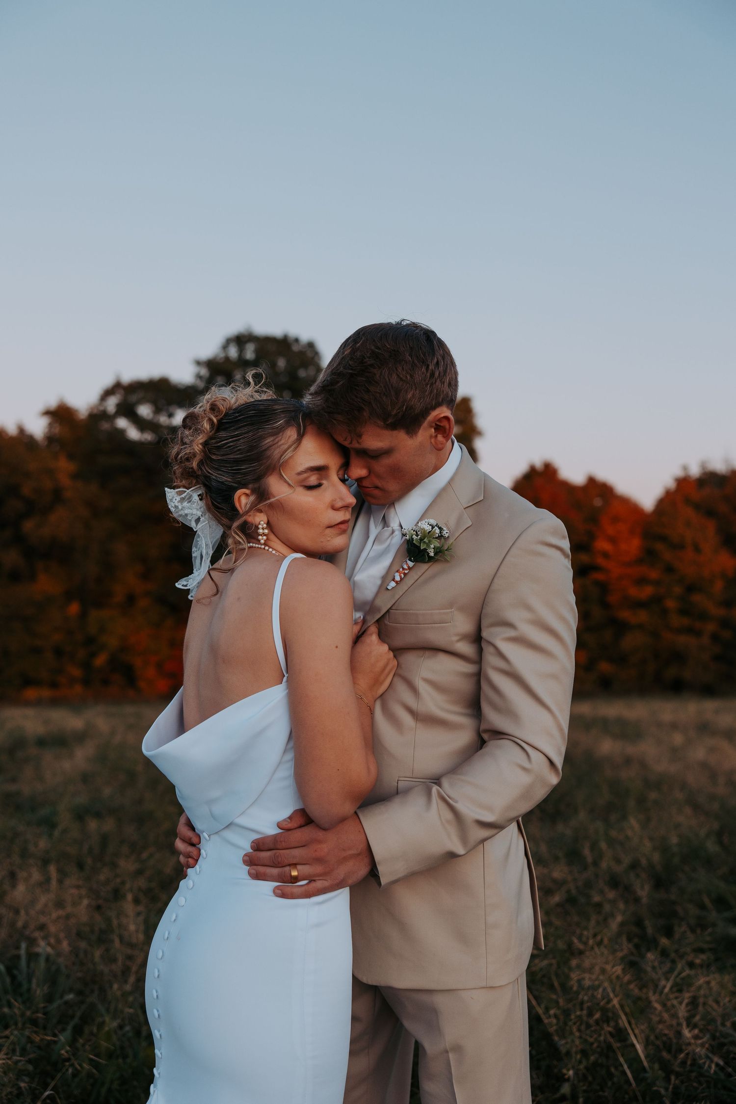 Newlyweds embrace in a field during golden hour with fall colors behind them.