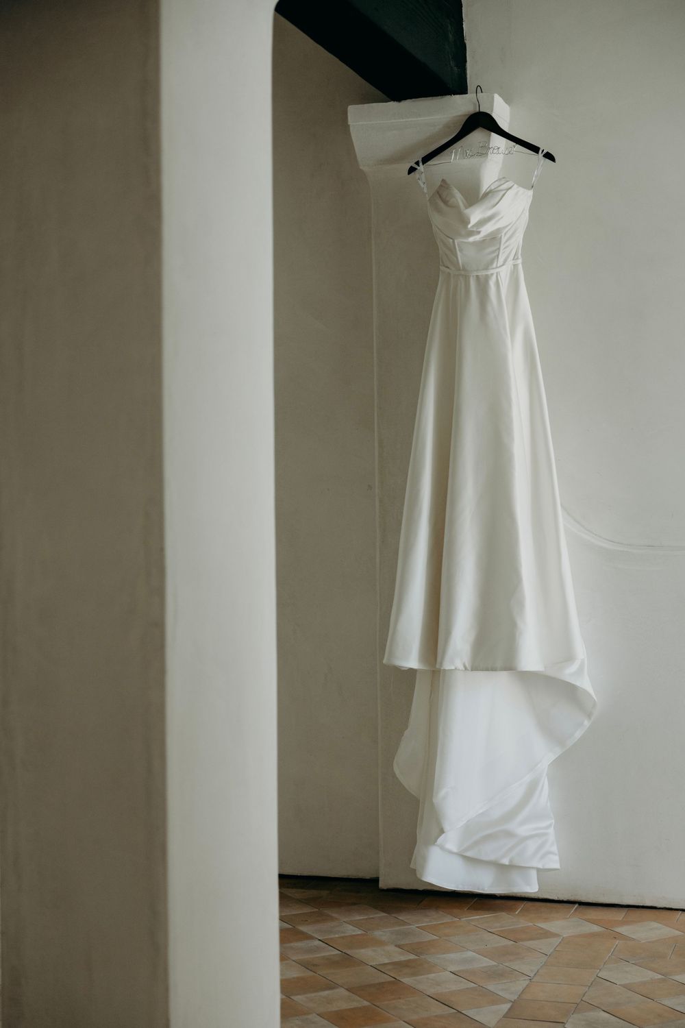 White wedding dress hanging against white wall in natural light.